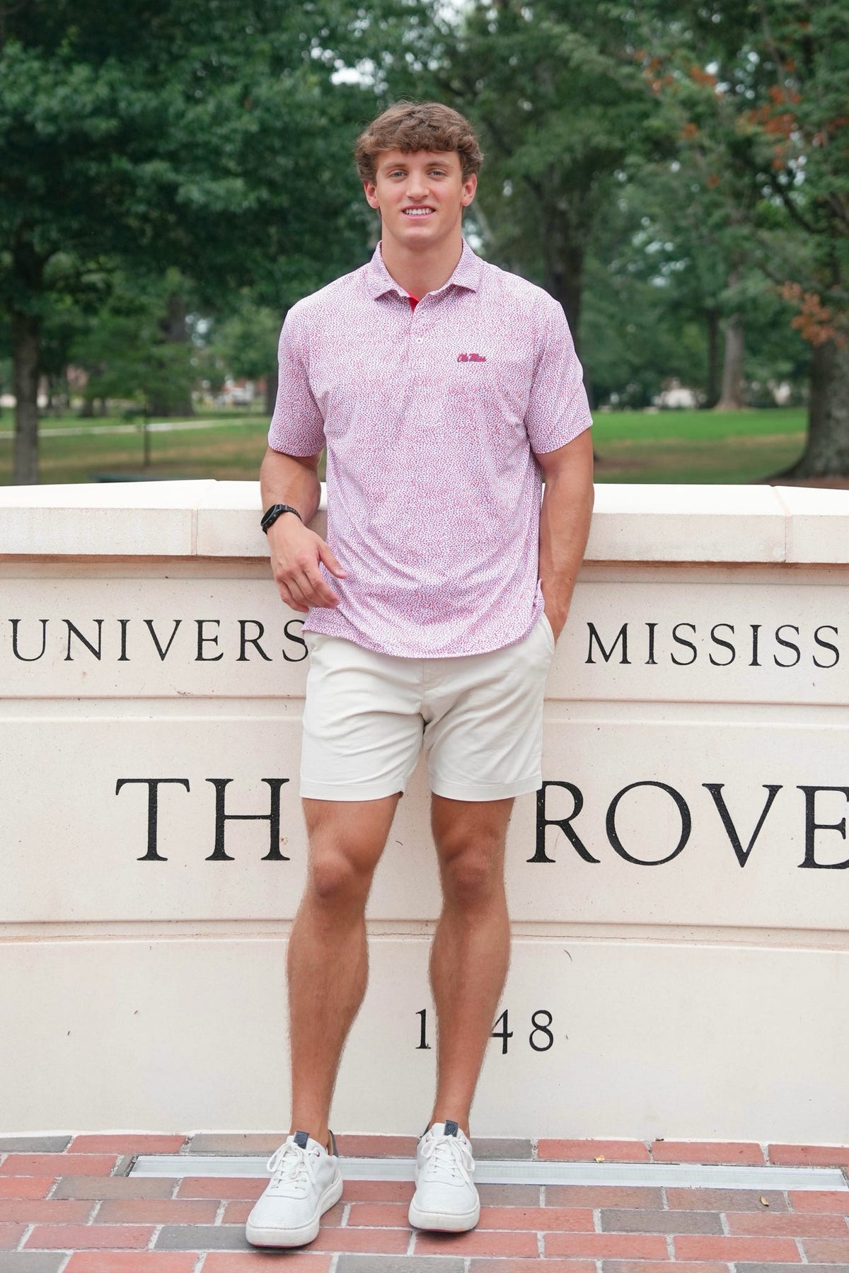 A young man stands outdoors in front of a stone wall with 'The Rovey' and 'University of Mississippi' text, wearing a light pink patterned polo shirt, beige shorts, and white sneakers.