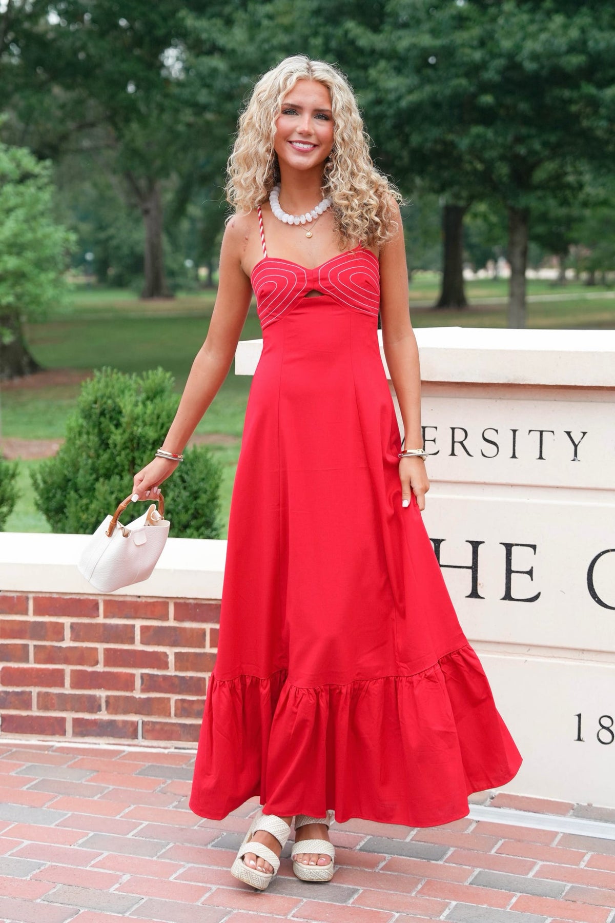 A woman stands outdoors wearing a bright red strapless maxi dress with a sweetheart neckline, ruched bodice, and a full tiered skirt. She is holding a small white handbag and wearing white platform sandals, with trees and a brick sign in the background.