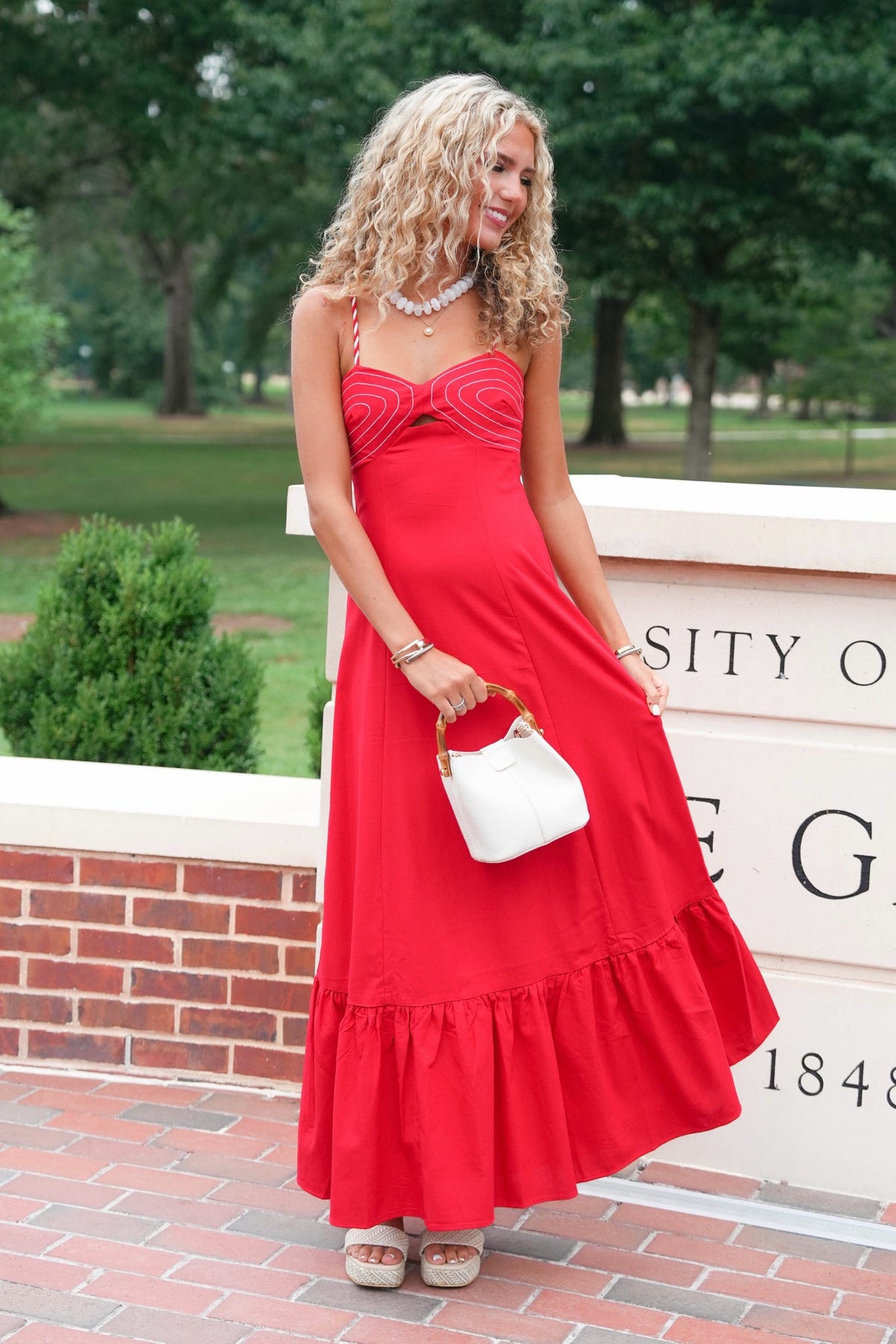 A woman wearing a bright red strapless maxi dress with a sweetheart neckline and ruffled hem, holding a small white handbag, standing outdoors in front of a brick wall and trees.
