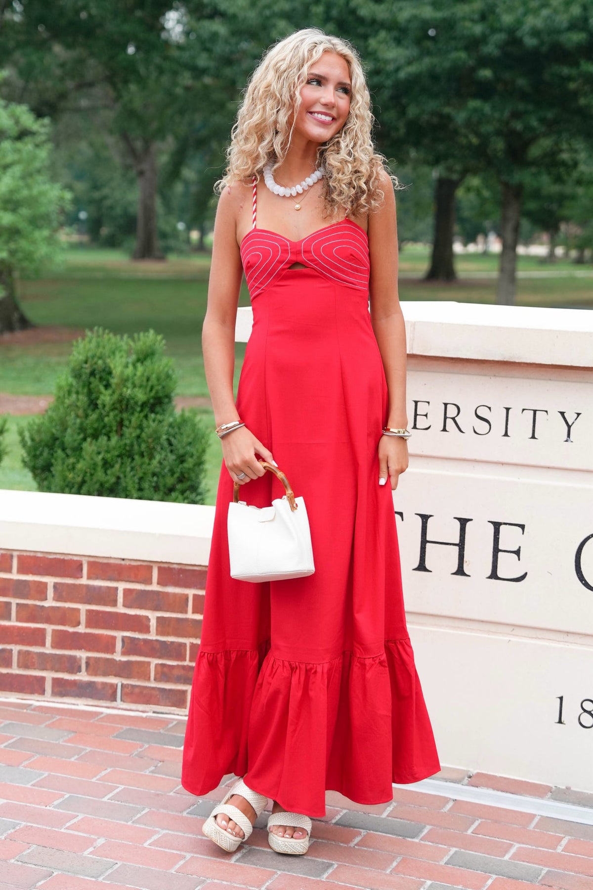 A woman stands outdoors wearing a bright red sleeveless maxi dress with a ruched bodice and flared hem. She is holding a small white handbag and wearing white platform sandals, with trees and a brick wall in the background.