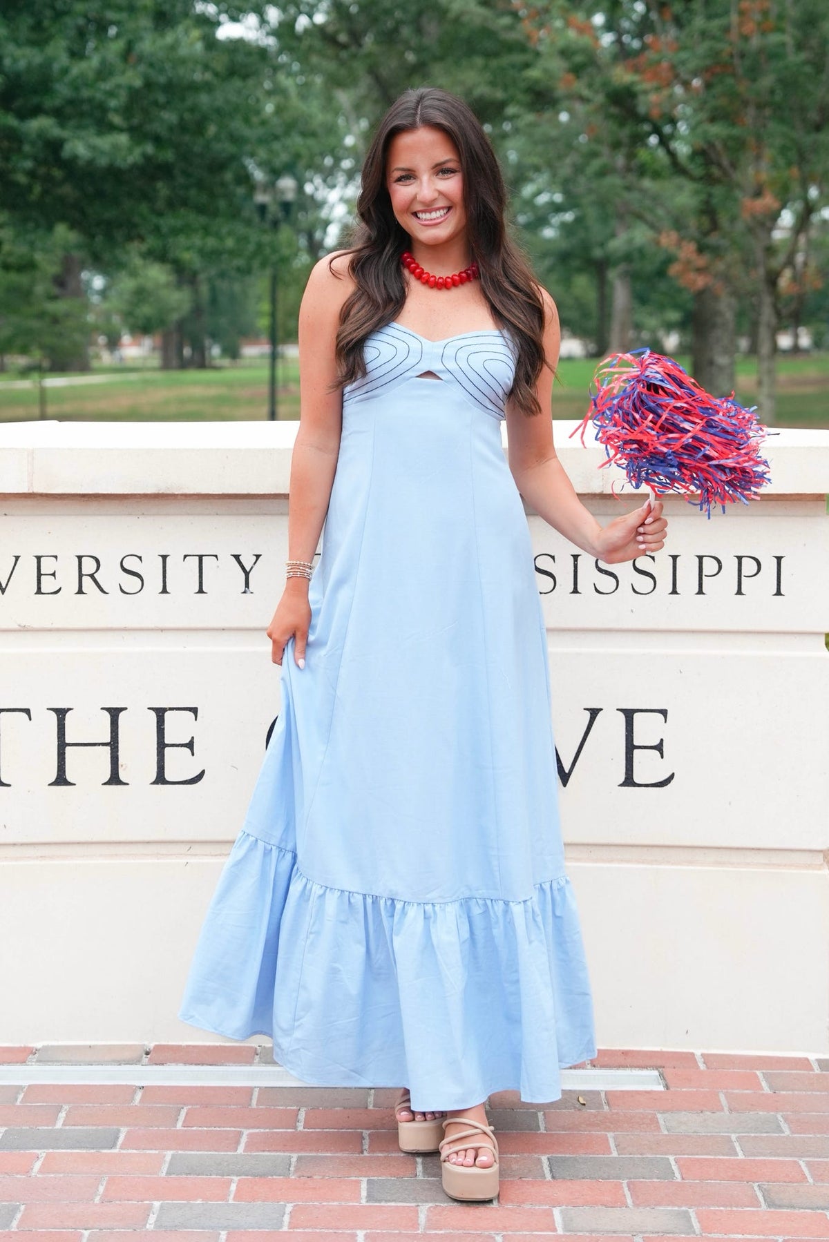 A woman stands outdoors wearing a light blue sleeveless maxi dress with a ruffled hem and a sweetheart neckline. She is holding a red and blue pom-pom in front of a brick walkway and a stone wall with university text.