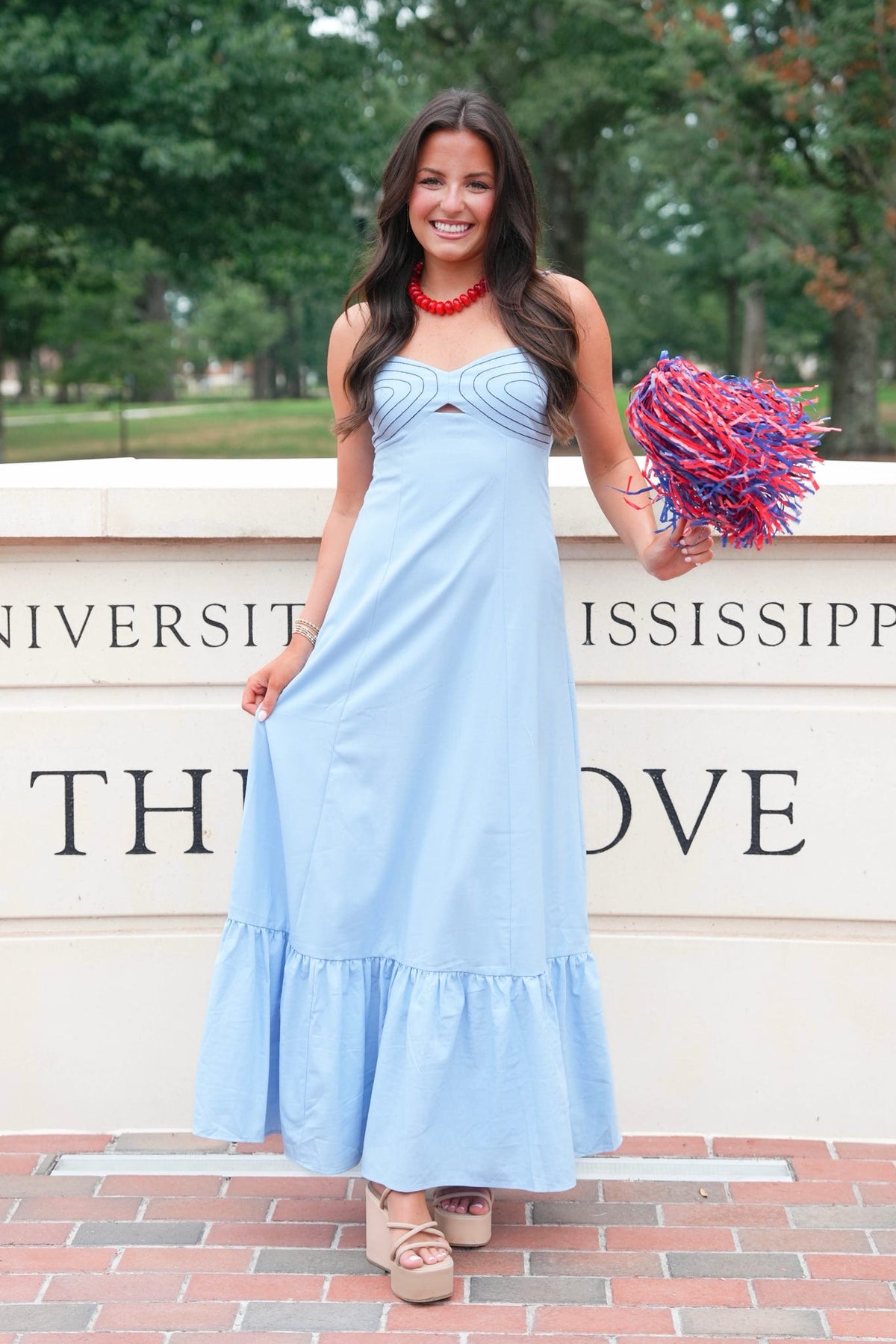 A woman stands outdoors in front of a stone wall with University of Mississippi text, wearing a light blue sleeveless maxi dress and holding a red and blue pom-pom.