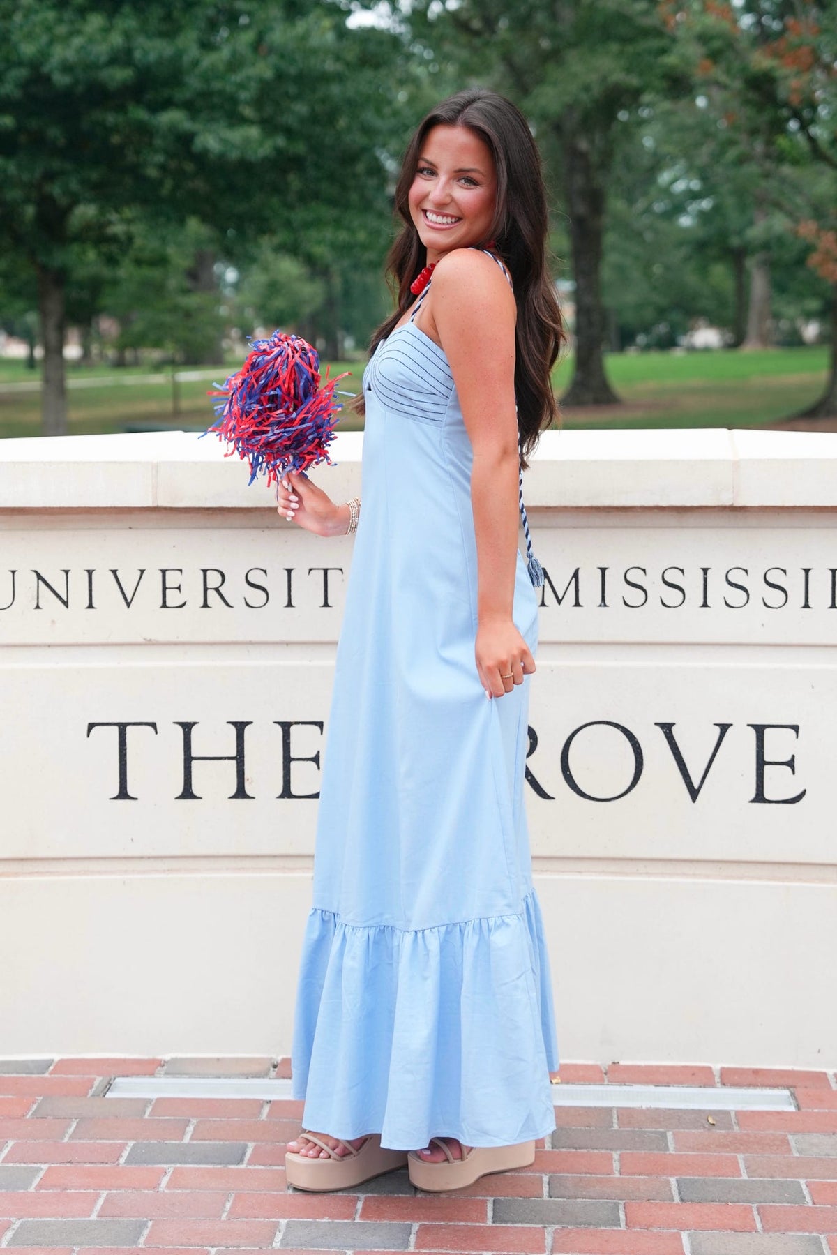 A woman wearing a light blue sleeveless maxi dress stands outdoors in front of a stone wall with 'University of Mississippi The Rose' text, holding a purple and red pom-pom.