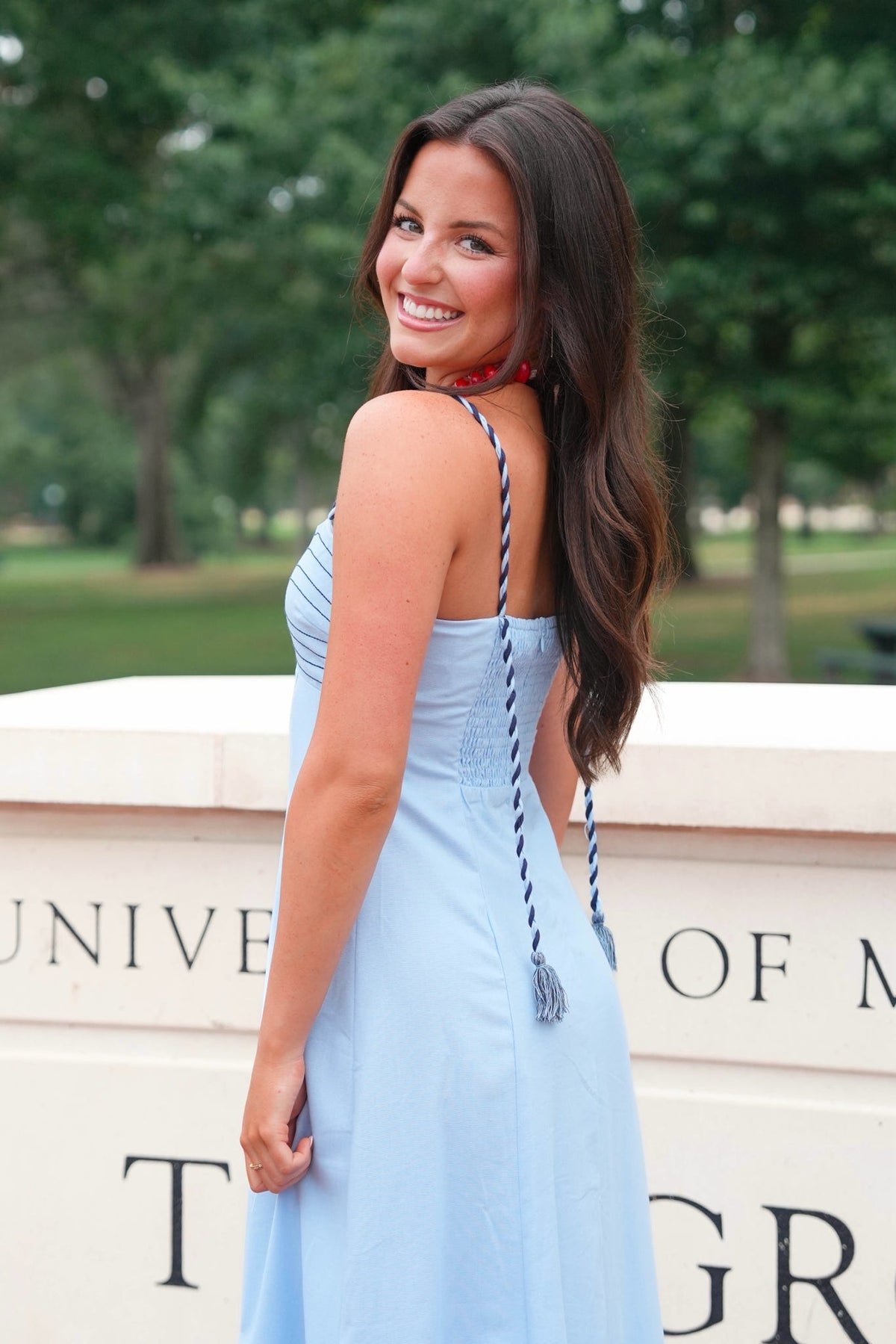 A woman wearing a light blue sleeveless maxi dress with thin braided straps and a red choker necklace, standing outdoors in front of a stone wall with trees in the background.