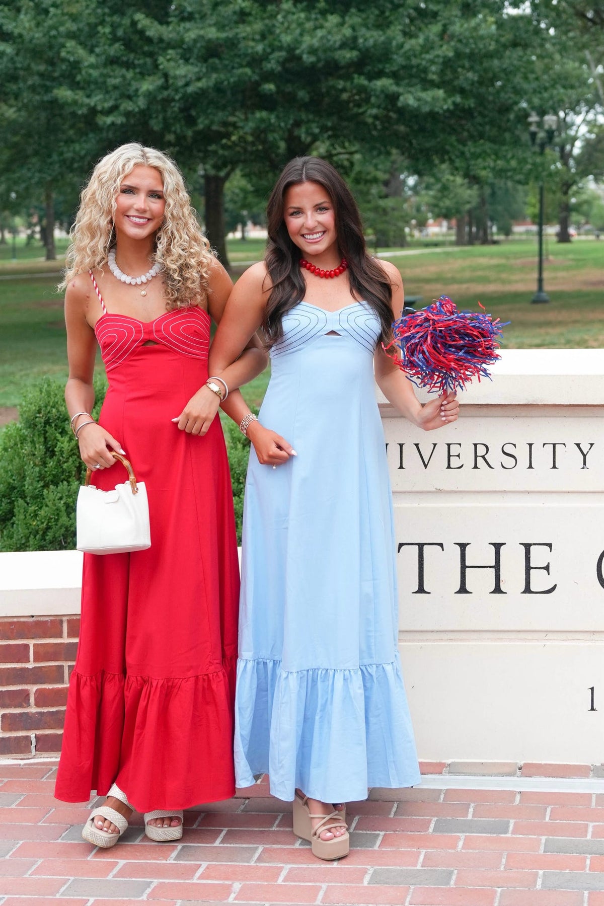 Two women standing outdoors in front of a brick wall and trees, wearing long strapless maxi dresses in red and light blue, each holding a matching pom-pom.