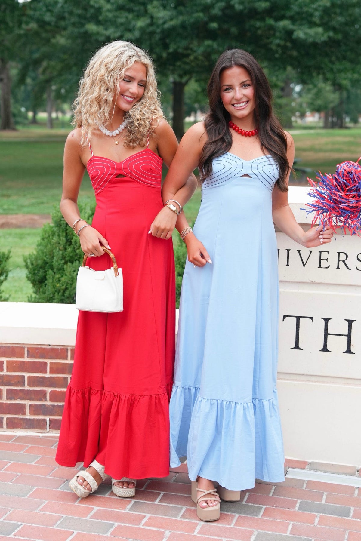 Two women standing outdoors in strapless maxi dresses, one in bright red and one in light blue, posing in front of a brick wall and trees.
