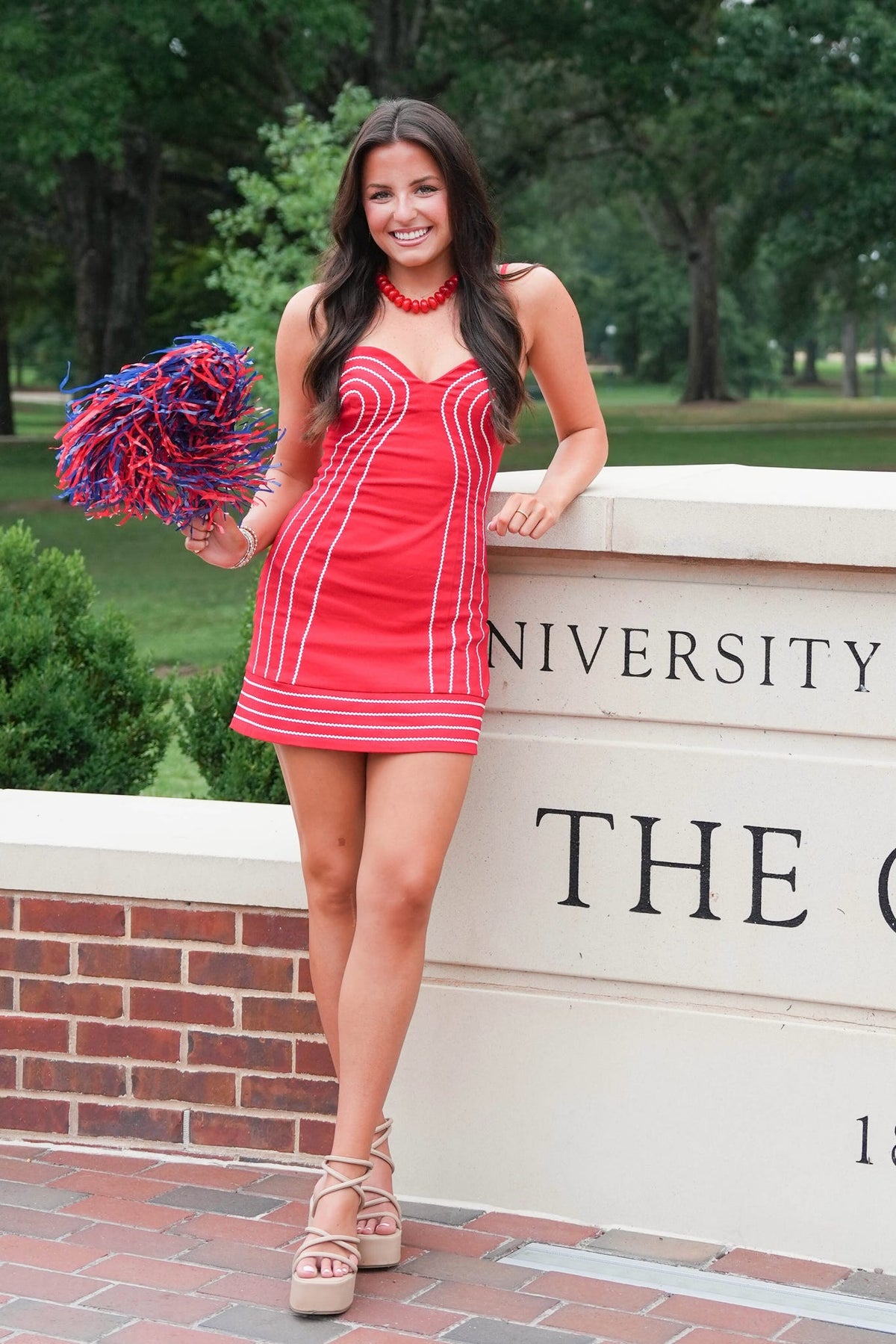 A woman posing outdoors in a red mini dress with white vertical stripes, holding a purple and red pom-pom. She is standing in front of a brick and concrete sign with trees in the background.