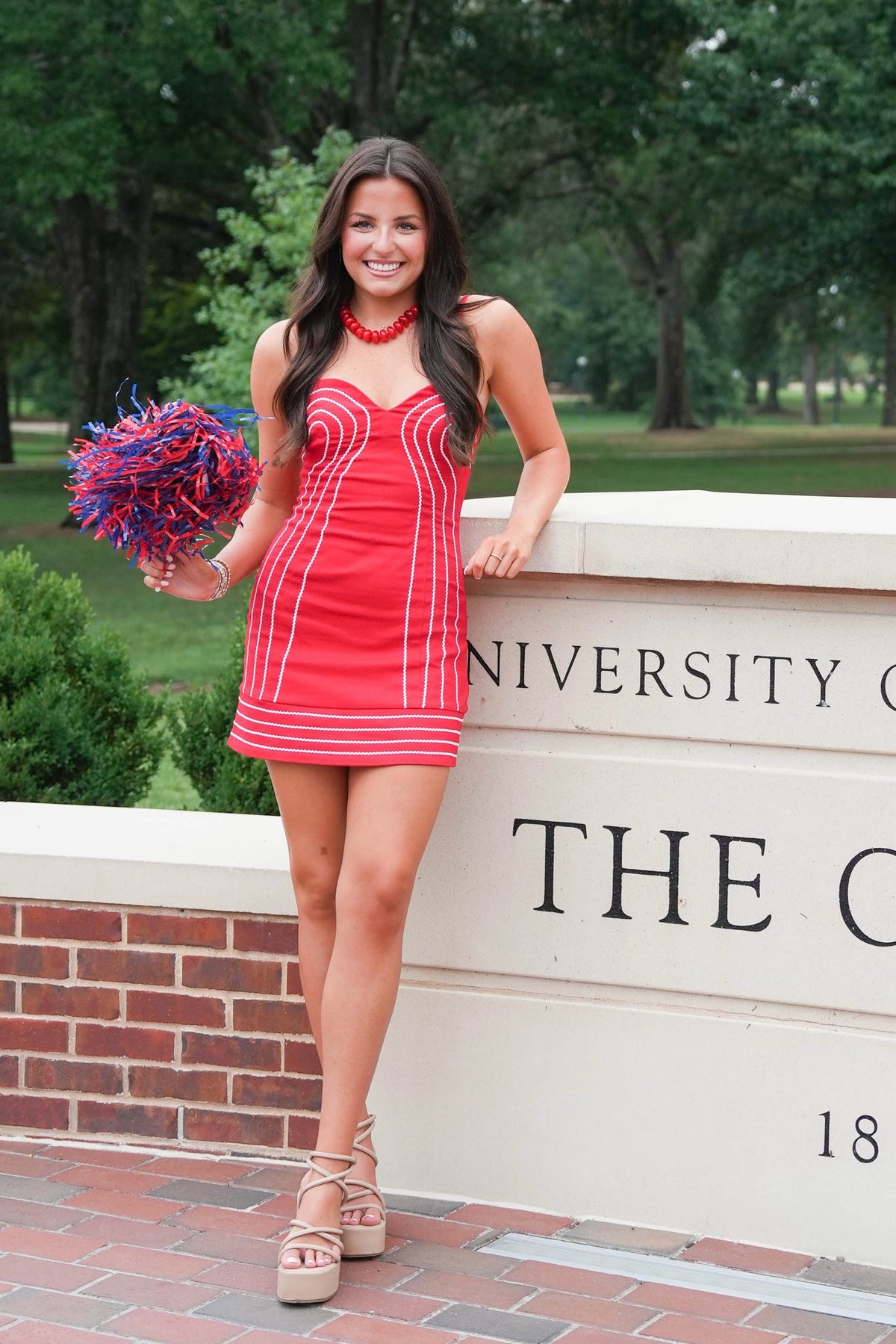 A woman posing outdoors in front of a brick and stone university sign, wearing a bright red mini dress with white vertical stripes and holding a blue and red pom-pom.