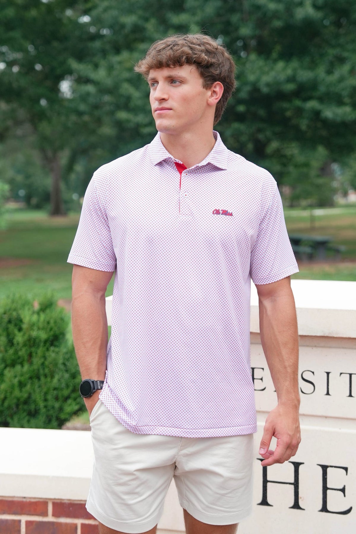 A man wearing a light purple and white micro-patterned polo shirt with a red collar and a red 'U O. Kent State' logo on the chest, paired with beige shorts, standing outdoors in front of a stone wall with green trees in the background.