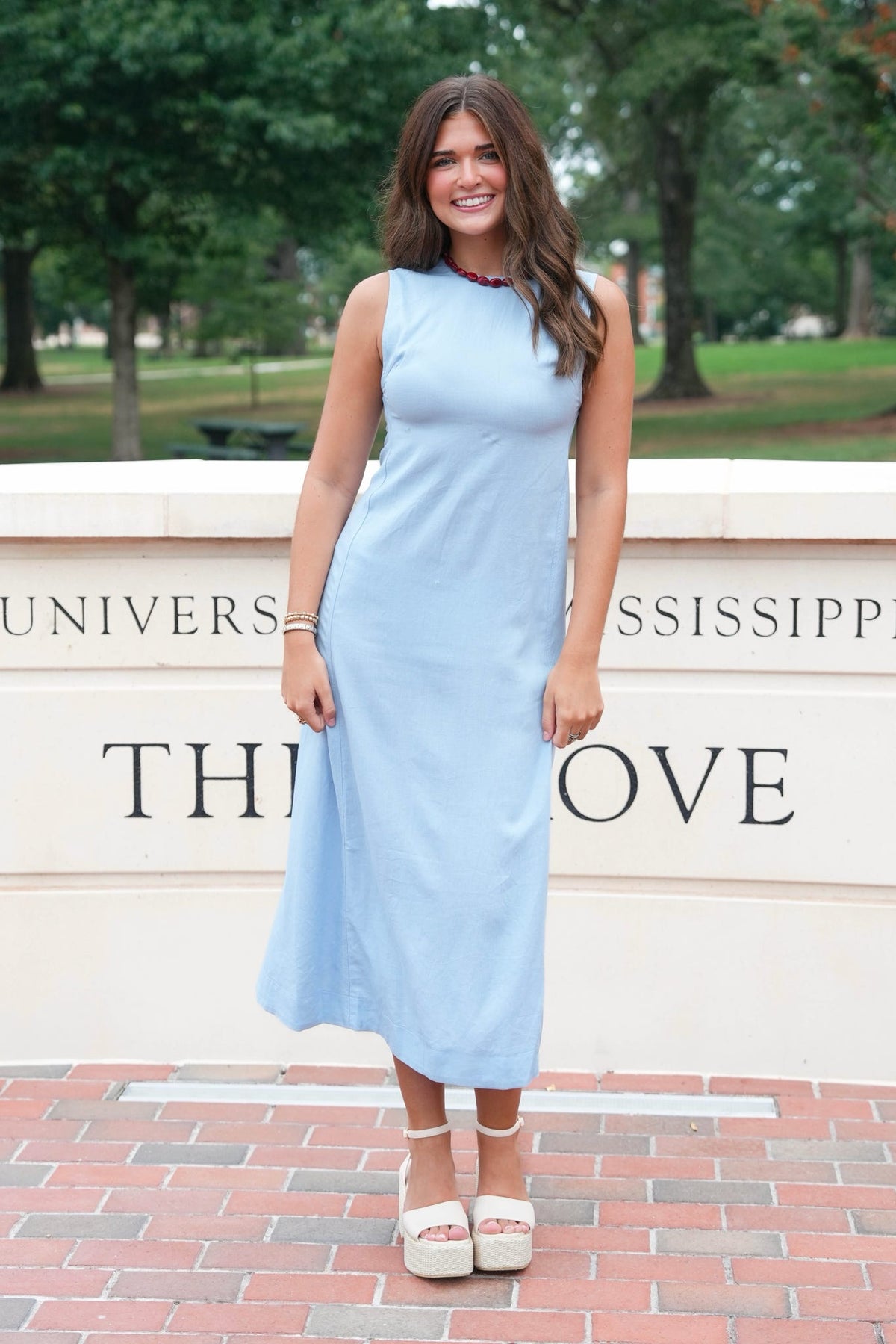 A woman stands outdoors wearing a sleeveless light blue midi dress and white platform sandals, posing in front of a stone wall with University of Mississippi lettering and trees in the background.