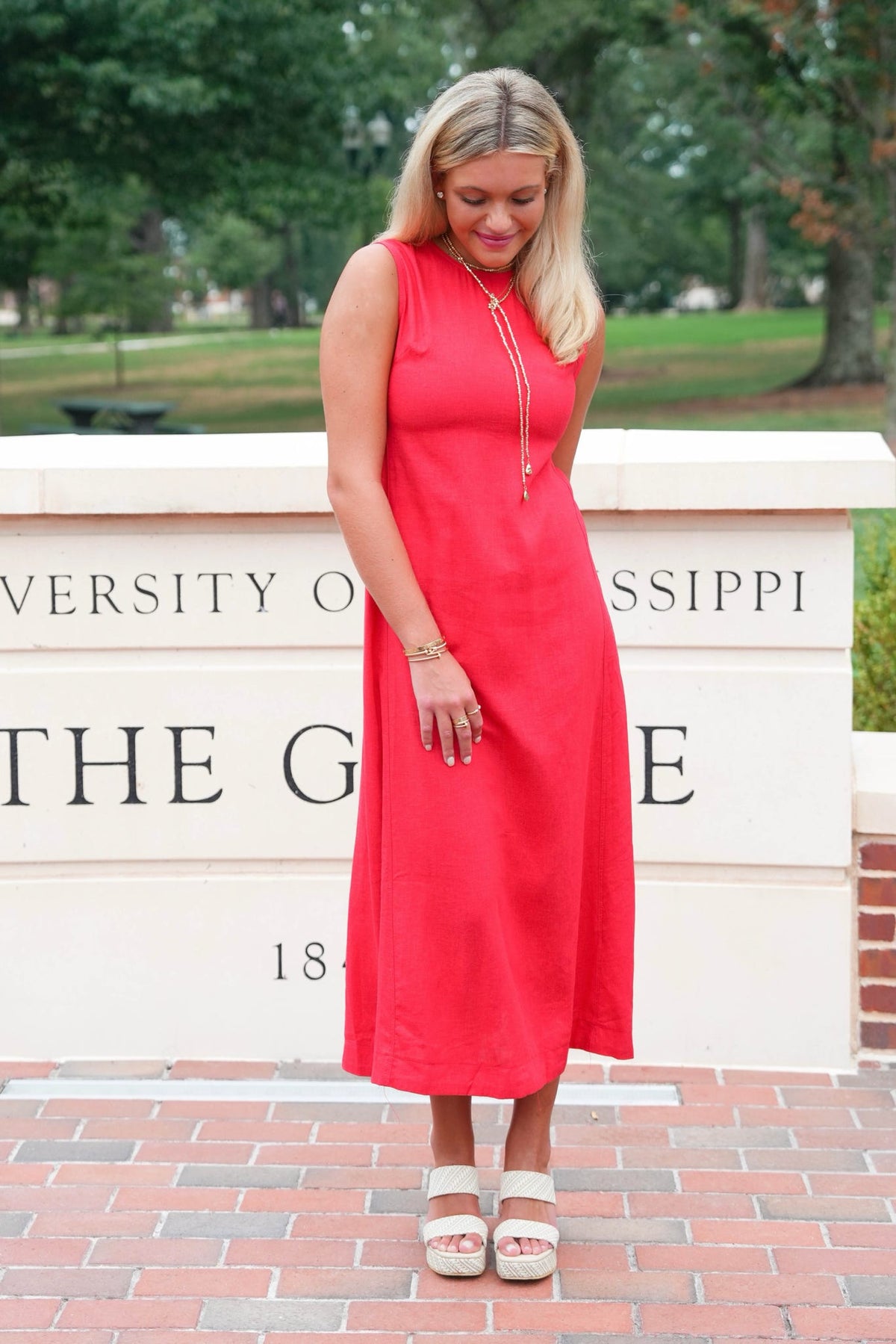 A woman wearing a sleeveless coral red midi dress and white platform sandals stands outdoors in front of a brick walkway and a sign that reads University of Mississippi.