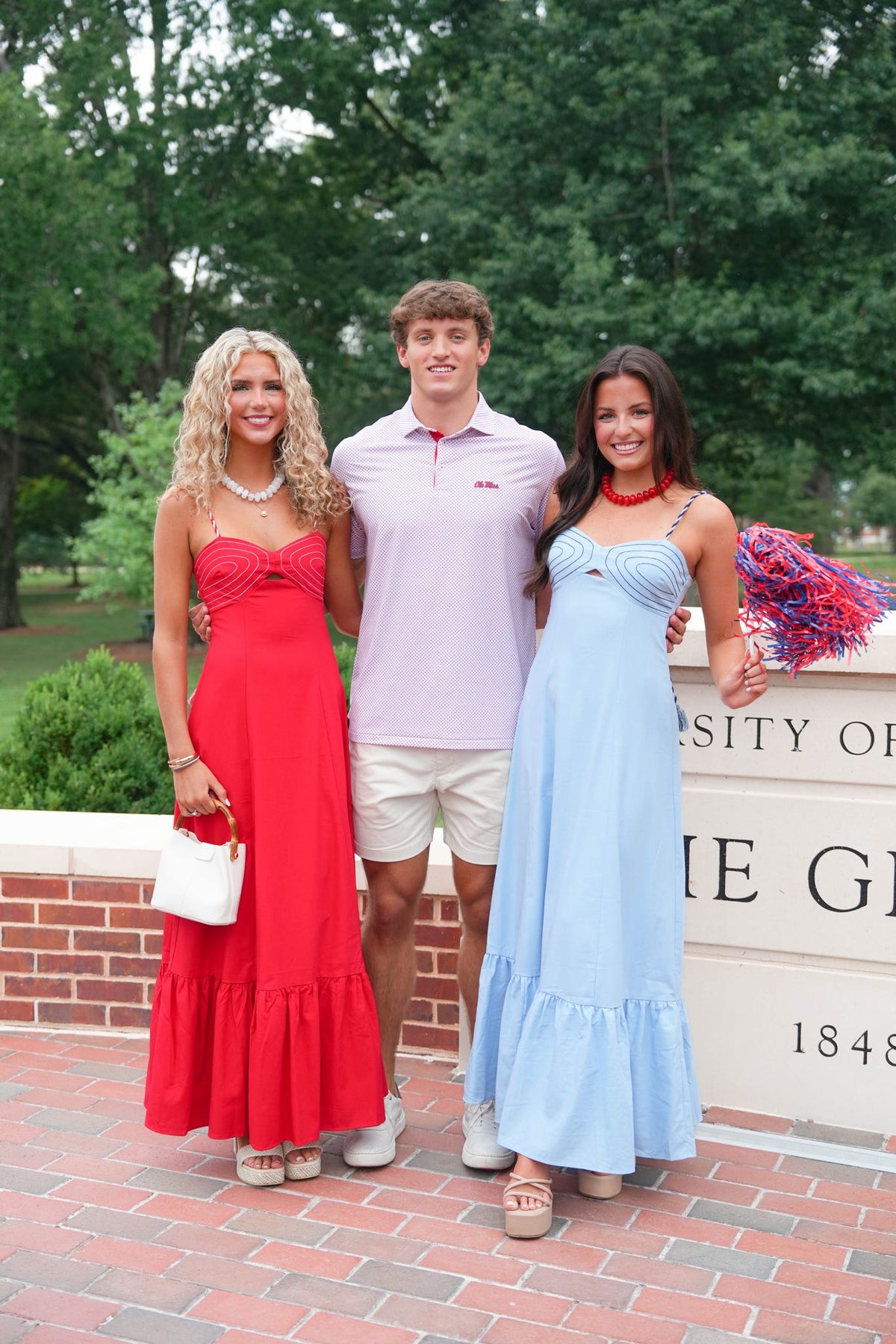 Three young adults posing outdoors in front of a brick wall and trees. The woman on the left wears a bright red strapless maxi dress and holds a white handbag.