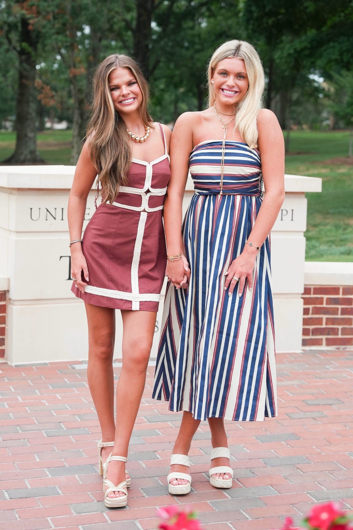 Two women standing outdoors on a brick walkway in front of a stone wall, wearing sleeveless dresses. The woman on the left wears a short burgundy dress with white trim and straps, and the woman on the right wears a strapless midi dress with vertical navy, white, and red stripes.