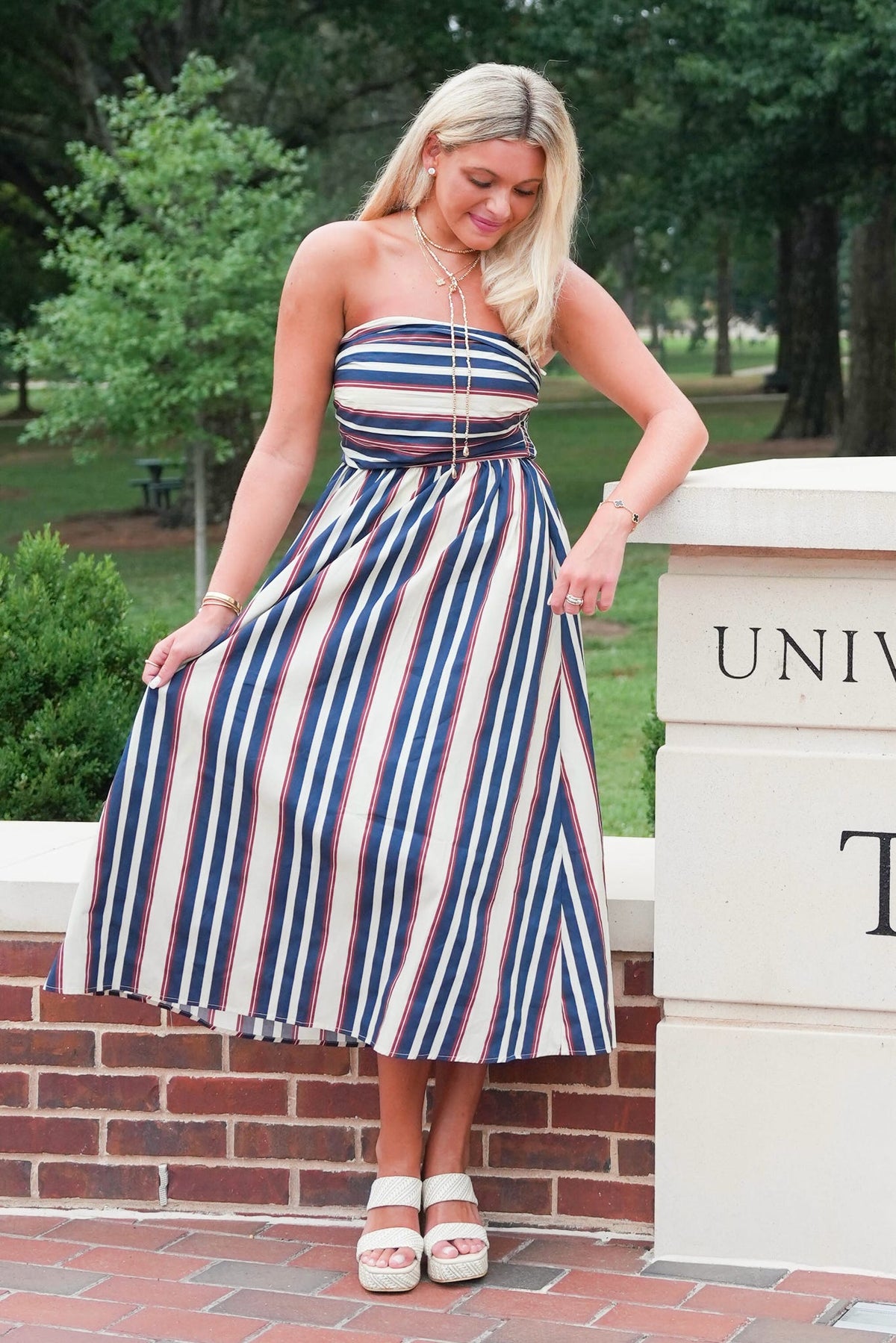 A woman wearing a strapless midi dress with vertical navy, red, and white stripes, posing outdoors next to a brick wall and a stone pillar.