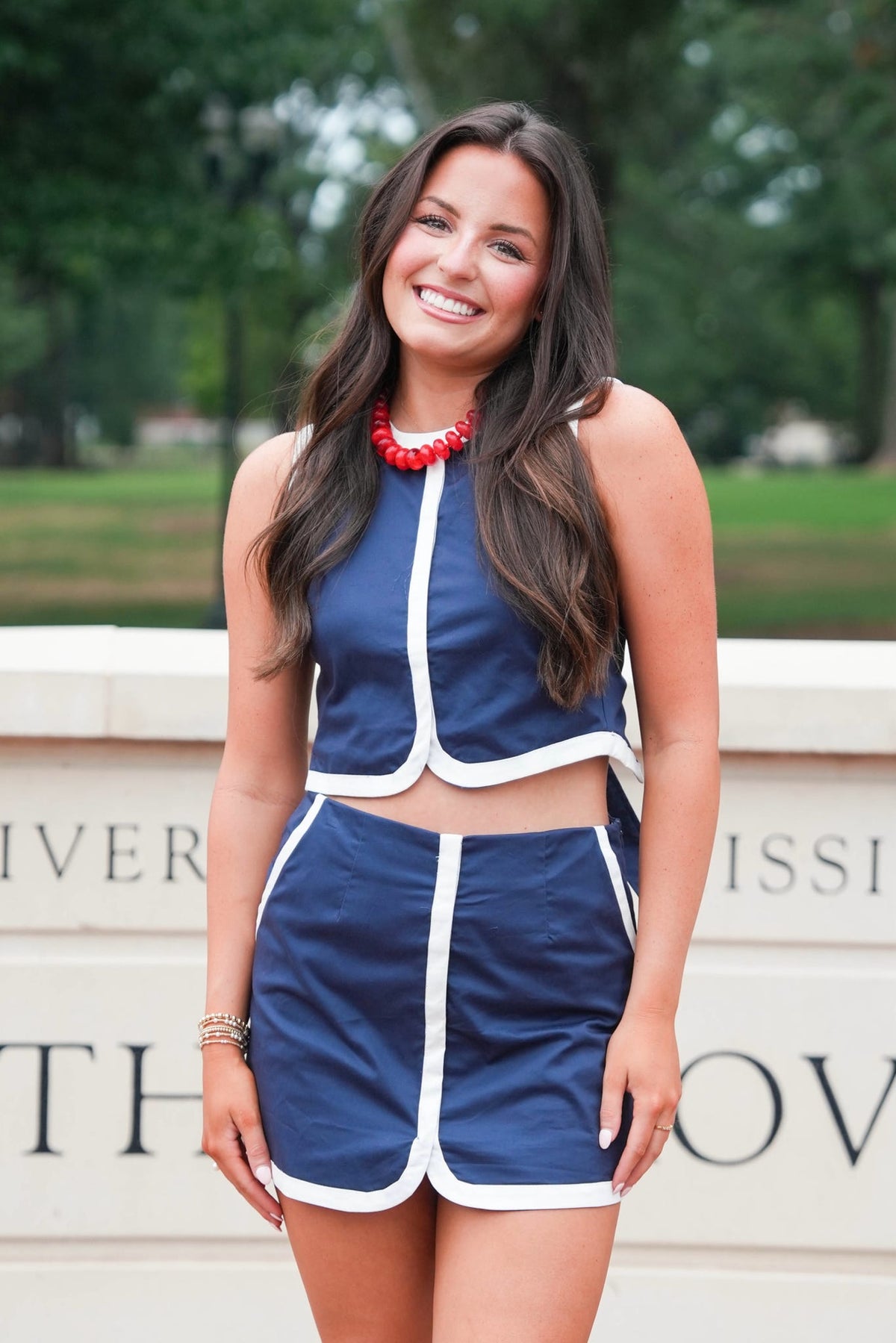 A woman smiling and wearing a navy blue two-piece athletic outfit with white piping, consisting of a sleeveless crop top and matching shorts, standing outdoors in front of a stone wall with green trees in the background.