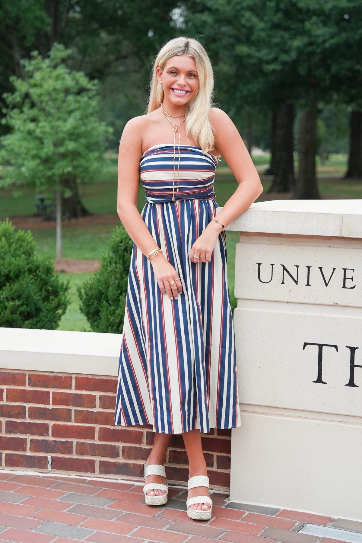 A woman stands outdoors wearing a strapless midi dress with vertical navy, red, and white stripes, leaning against a white brick and stone wall with trees in the background.