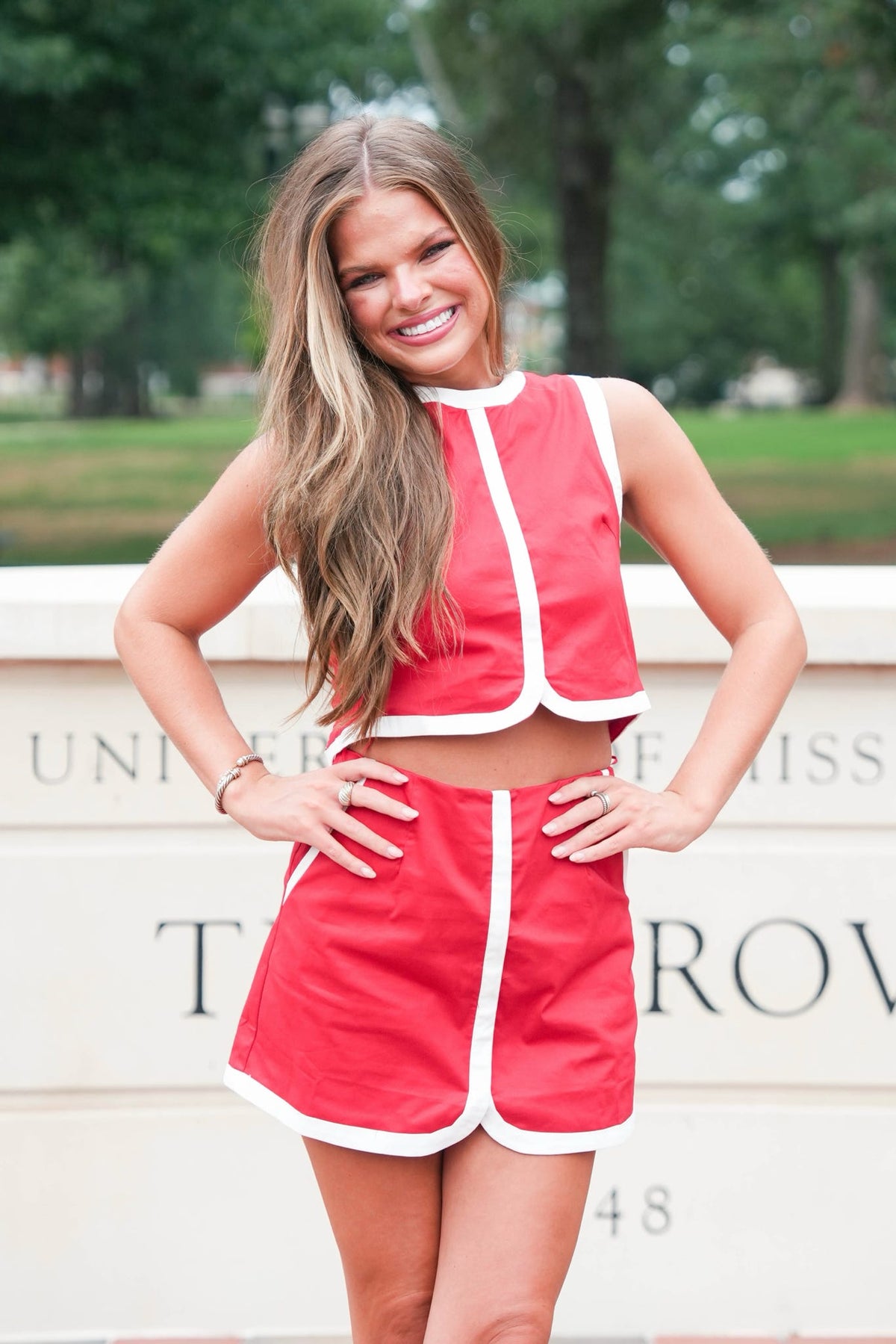 A smiling young woman stands outdoors wearing a bright red two-piece outfit with white trim, consisting of a sleeveless cropped top and matching shorts. The background shows a light-colored stone wall with university text and blurred green trees.