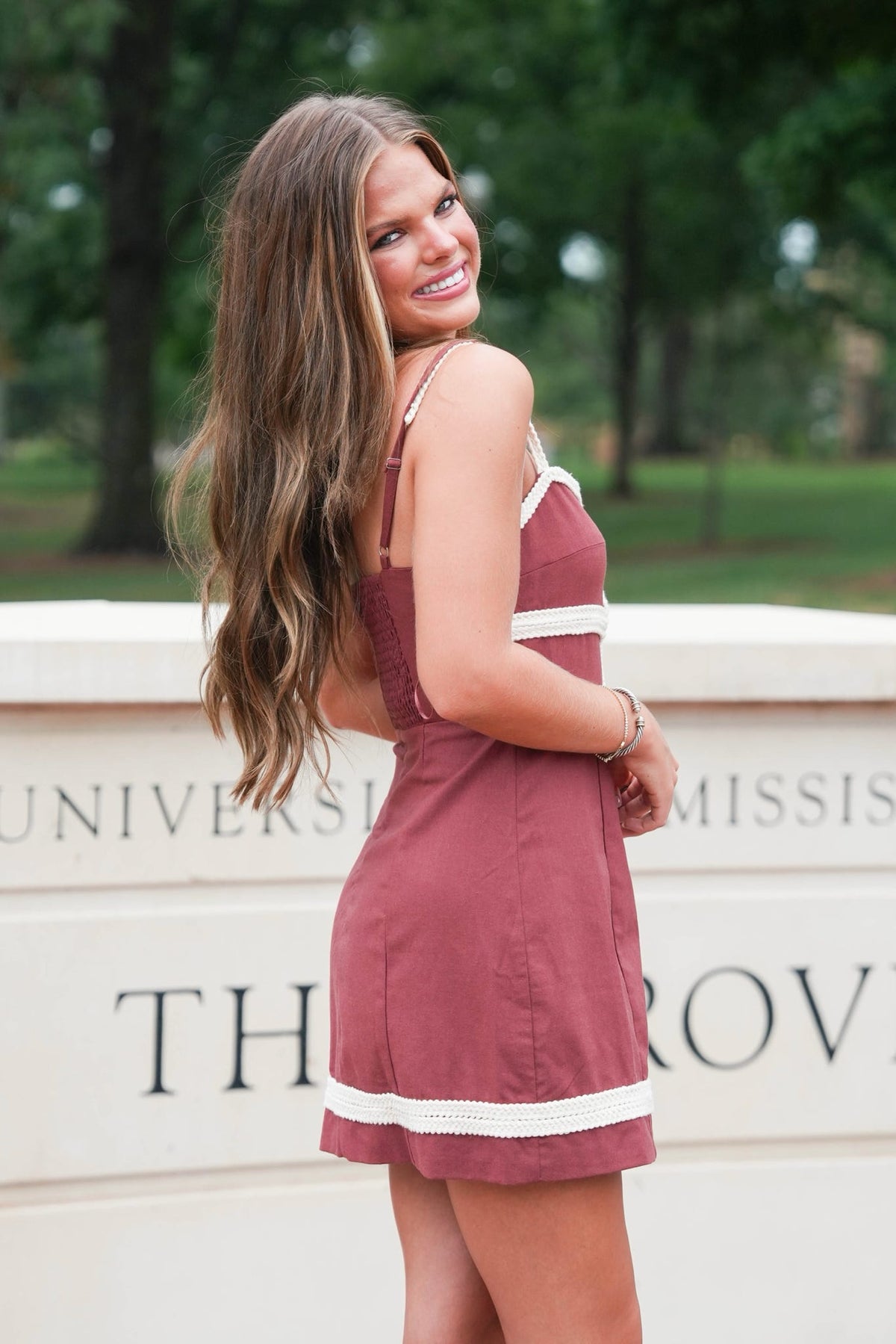 A woman wearing a dusty rose mini dress with white trim, standing outdoors in front of a stone wall with 'University of Tennessee' text in the background.
