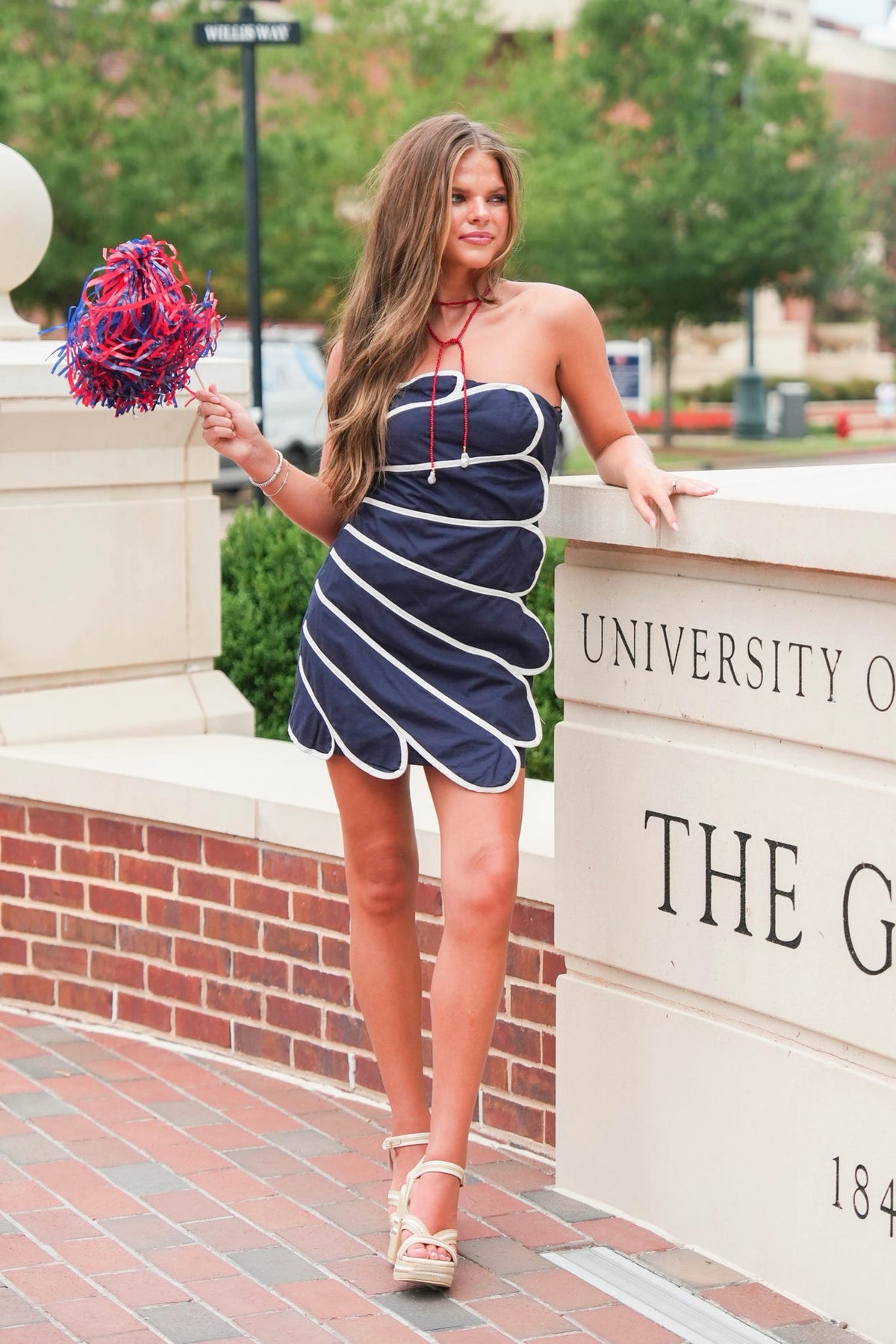 A woman posing outdoors in a navy blue strapless mini dress with white diagonal stripes and white piping. She is standing next to a brick and stone walkway with 'UNIVERSITY OF THE G' visible on the stone wall, holding a red and blue pom-pom.