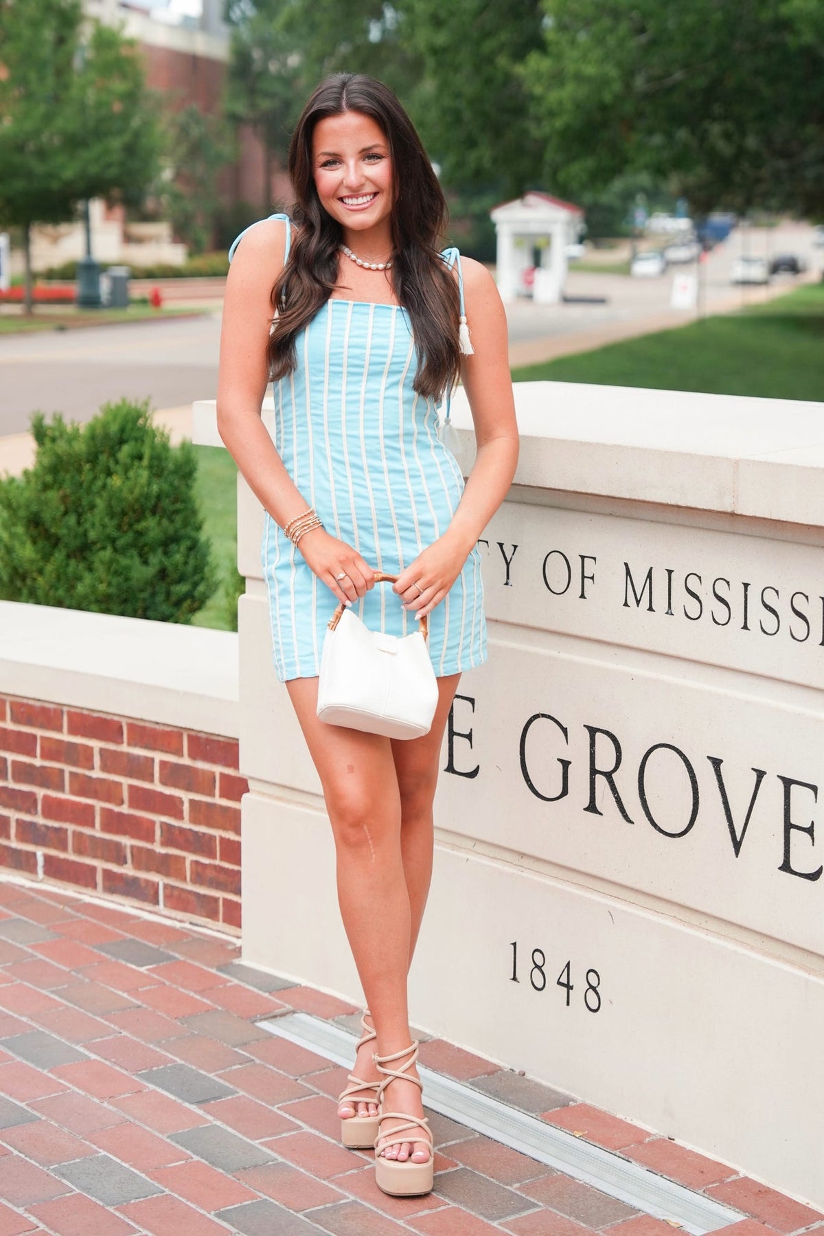 A woman stands outdoors in front of a stone sign reading 'University of Mississippi Grove 1848', wearing a light blue and white vertically striped sleeveless mini dress, holding a small white handbag, and wearing beige strappy platform sandals.