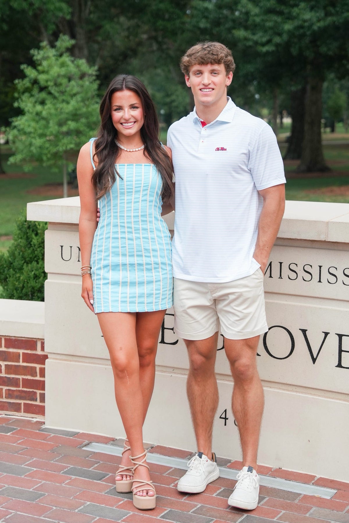 A young woman in a light blue and white striped mini dress and a man in a white polo shirt and beige shorts standing together outdoors in front of a brick and stone wall with green trees in the background.
