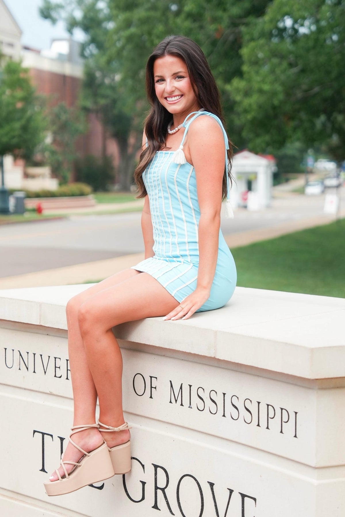 A young woman with long dark hair sits on a stone monument at the University of Mississippi The Grove, wearing a light blue and white striped mini dress and beige platform wedge sandals.