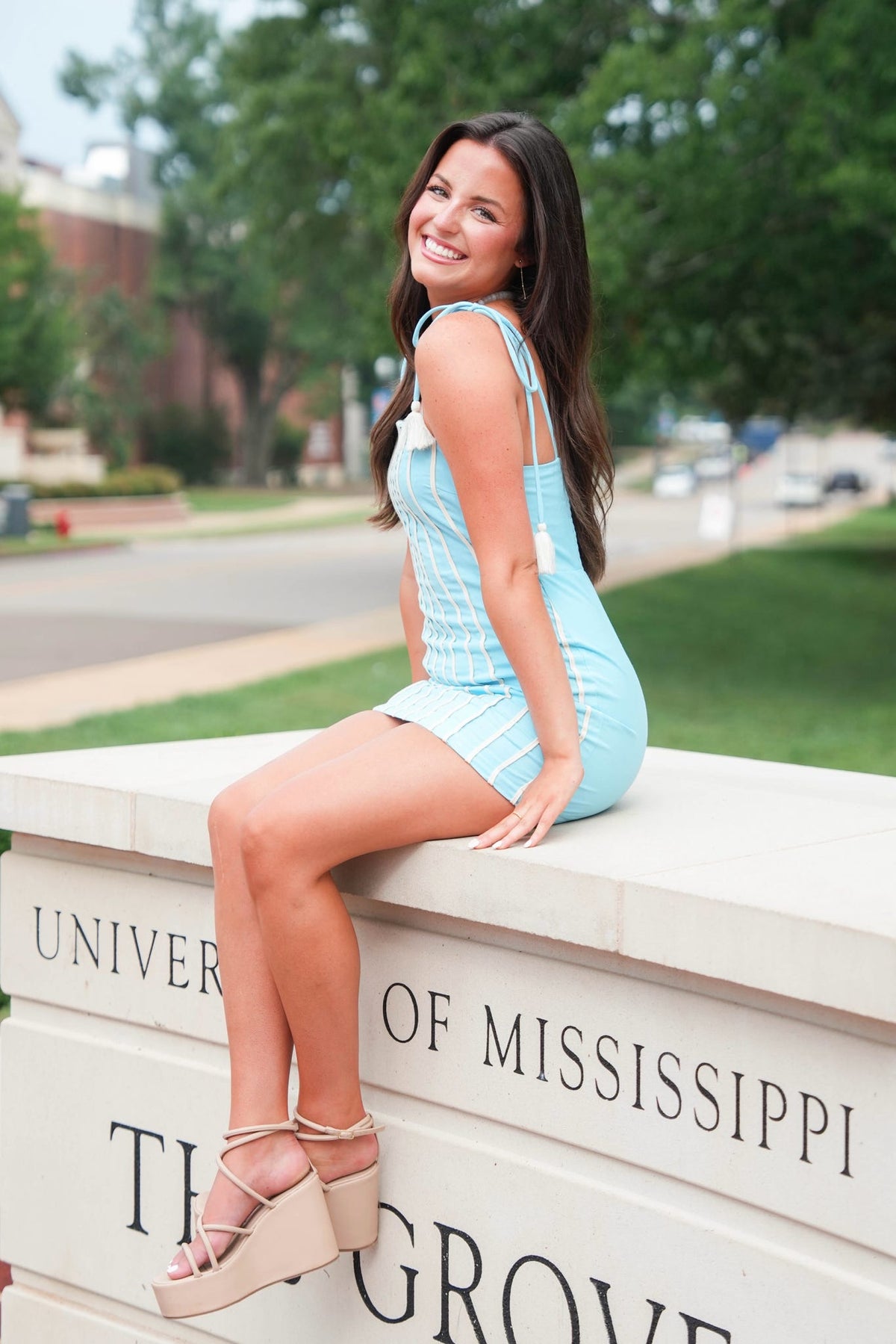 A woman sitting on a stone monument outdoors, wearing a light blue sleeveless mini dress and beige platform sandals. The background shows a blurred campus setting with trees and the University of Mississippi sign visible.