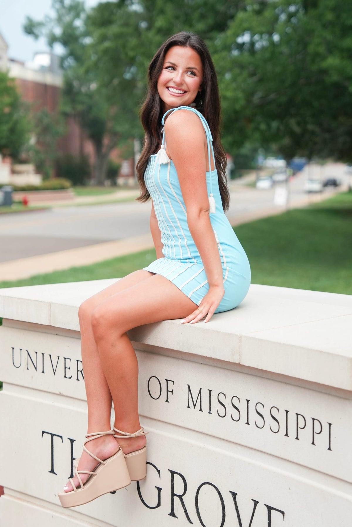 A woman sitting on a stone monument outdoors, wearing a light blue sleeveless mini dress and beige strappy platform sandals. The monument is inscribed with 'UNIVERSITY OF MISSISSIPPI' and 'THE GROVE'.