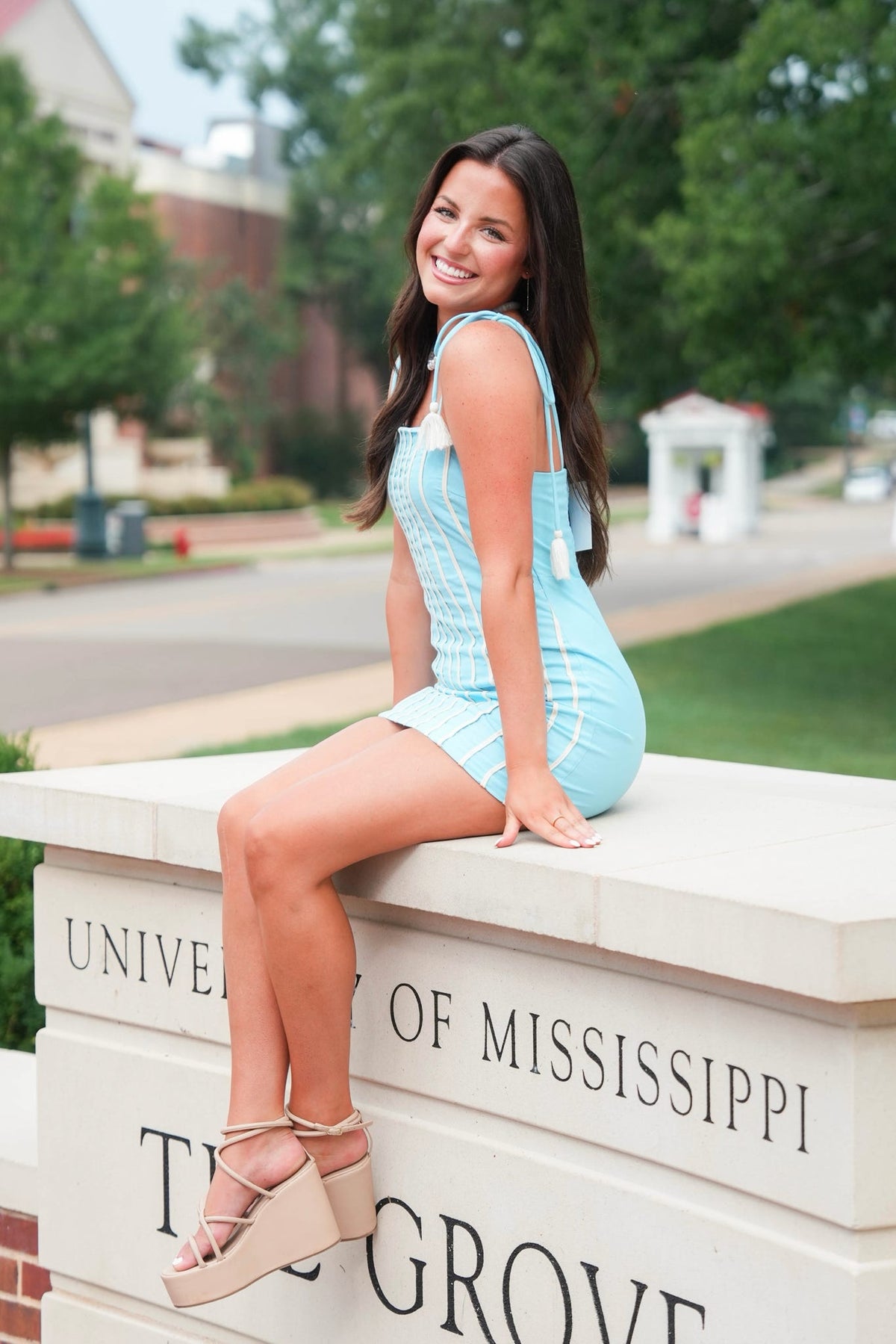 A young woman with long dark hair sits on a stone wall at the University of Mississippi Thurgood E. Brown Graduate Center, wearing a light blue sleeveless mini dress and beige platform sandals.