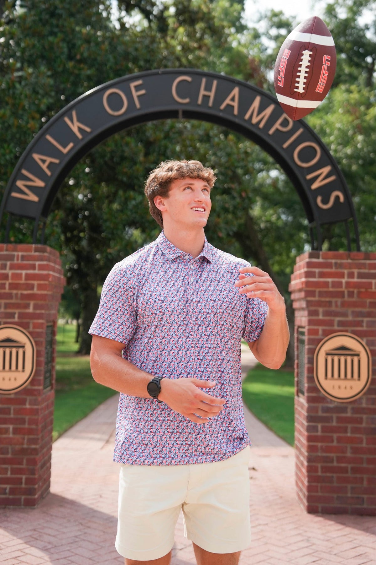 A young man stands outdoors in front of a brick arch reading 'WALK OF CHAMPIONS' while tossing a football. He is wearing a short-sleeve patterned button-up shirt in light purple and white, paired with cream shorts.