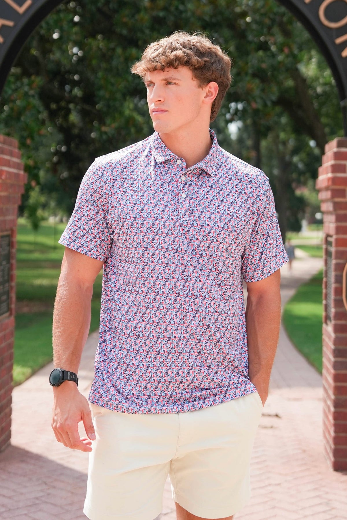A man wearing a short-sleeve button-up polo shirt with a dense red, purple, and white geometric pattern, paired with cream shorts, standing outdoors under a brick archway.