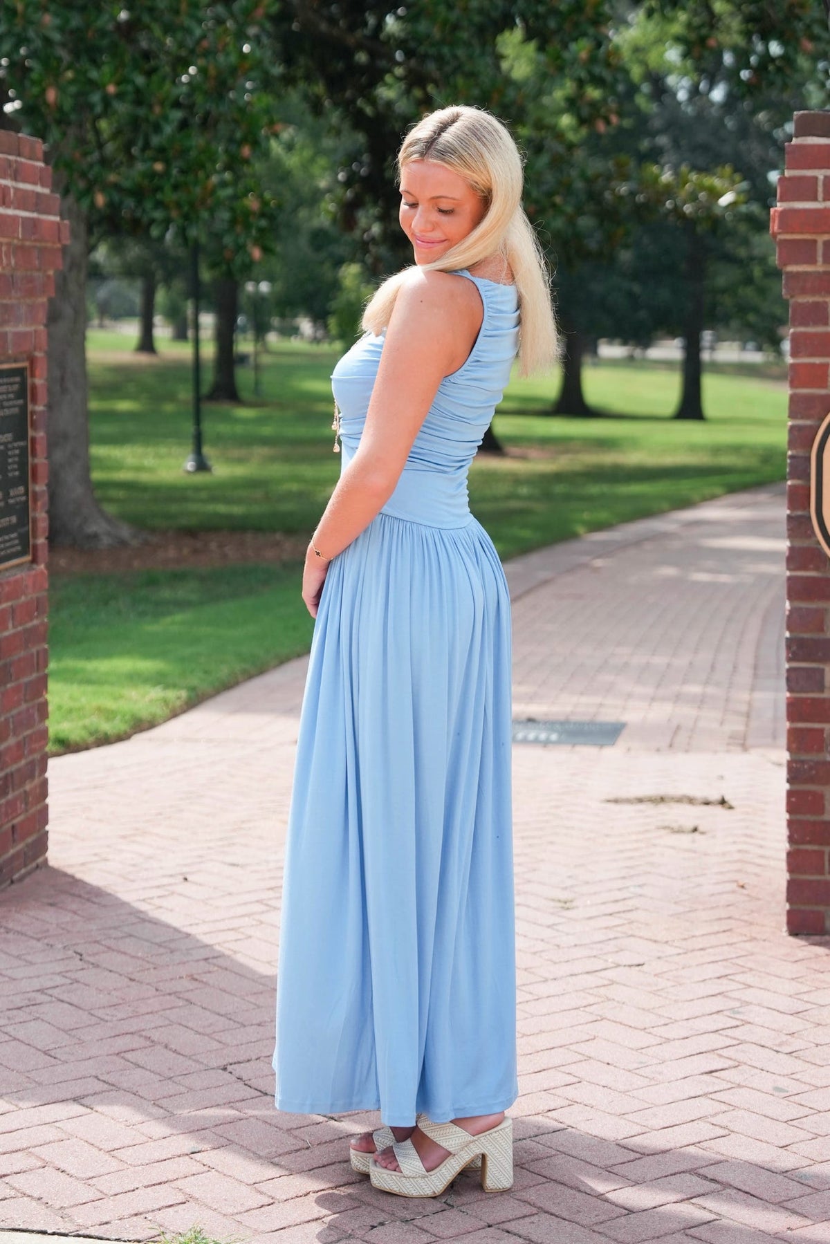 A woman wearing a light blue sleeveless maxi dress and white platform sandals stands on a brick pathway in a park setting.