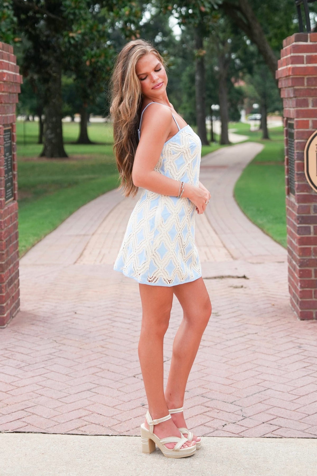 A woman stands on a brick pathway between brick pillars, wearing a light blue and white patterned mini dress with beige platform sandals in an outdoor park setting.