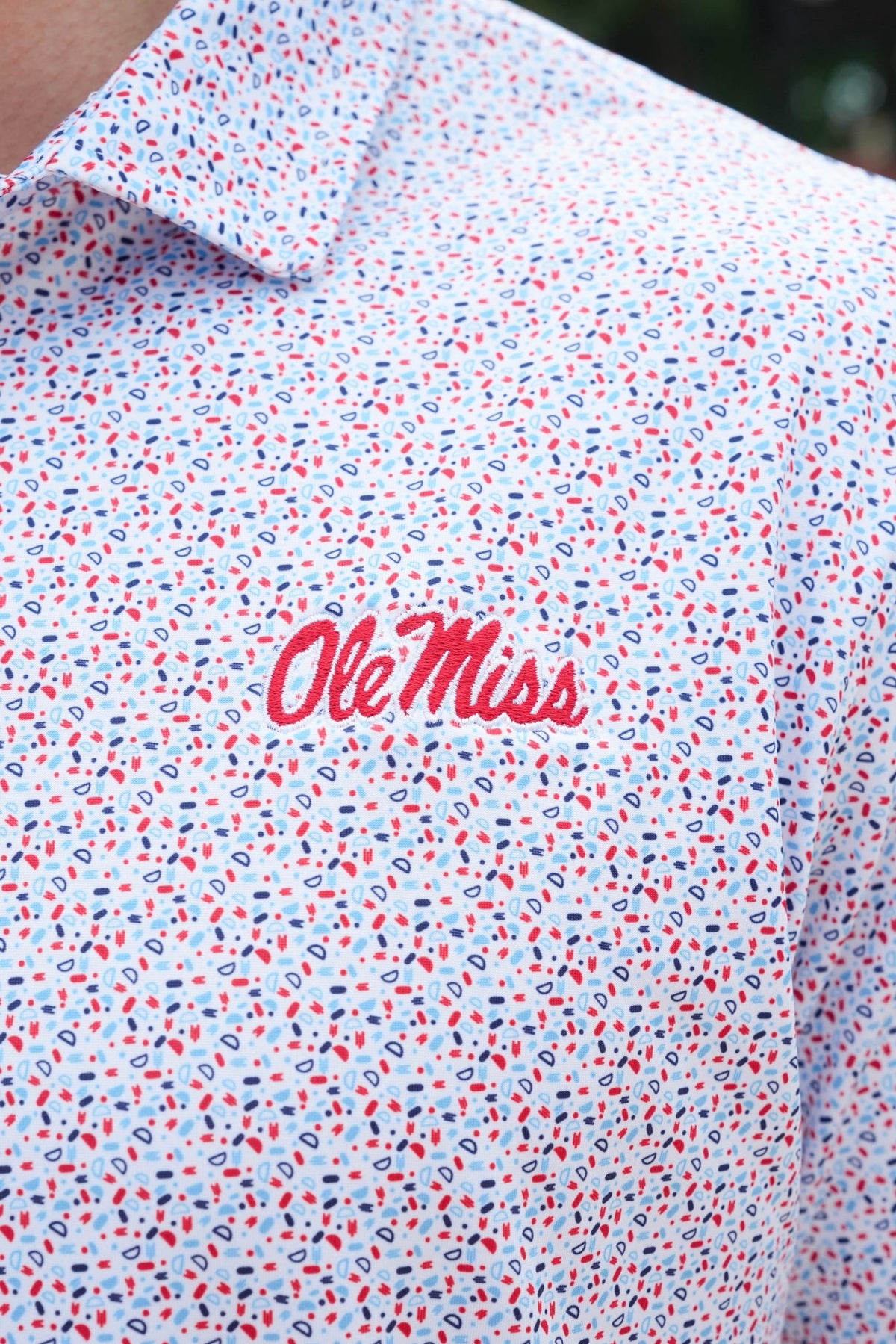 Close-up of a white short-sleeve button-up shirt with a dense red, blue, and black micro-pattern, featuring the red script 'OleMiss' logo on the chest.