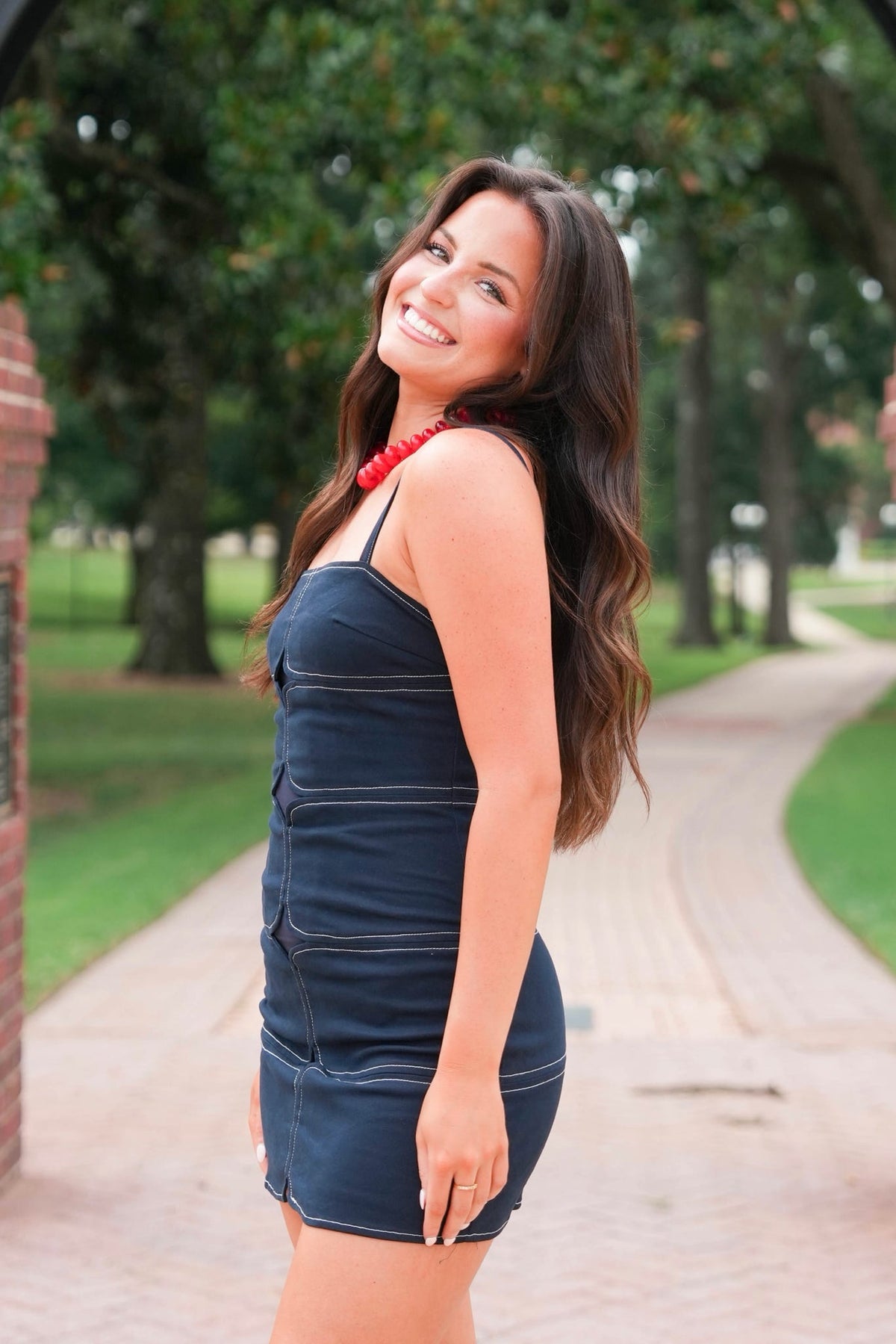 A woman with long brown hair stands outdoors on a brick pathway wearing a dark navy blue denim mini dress with thin straps and a red beaded necklace.