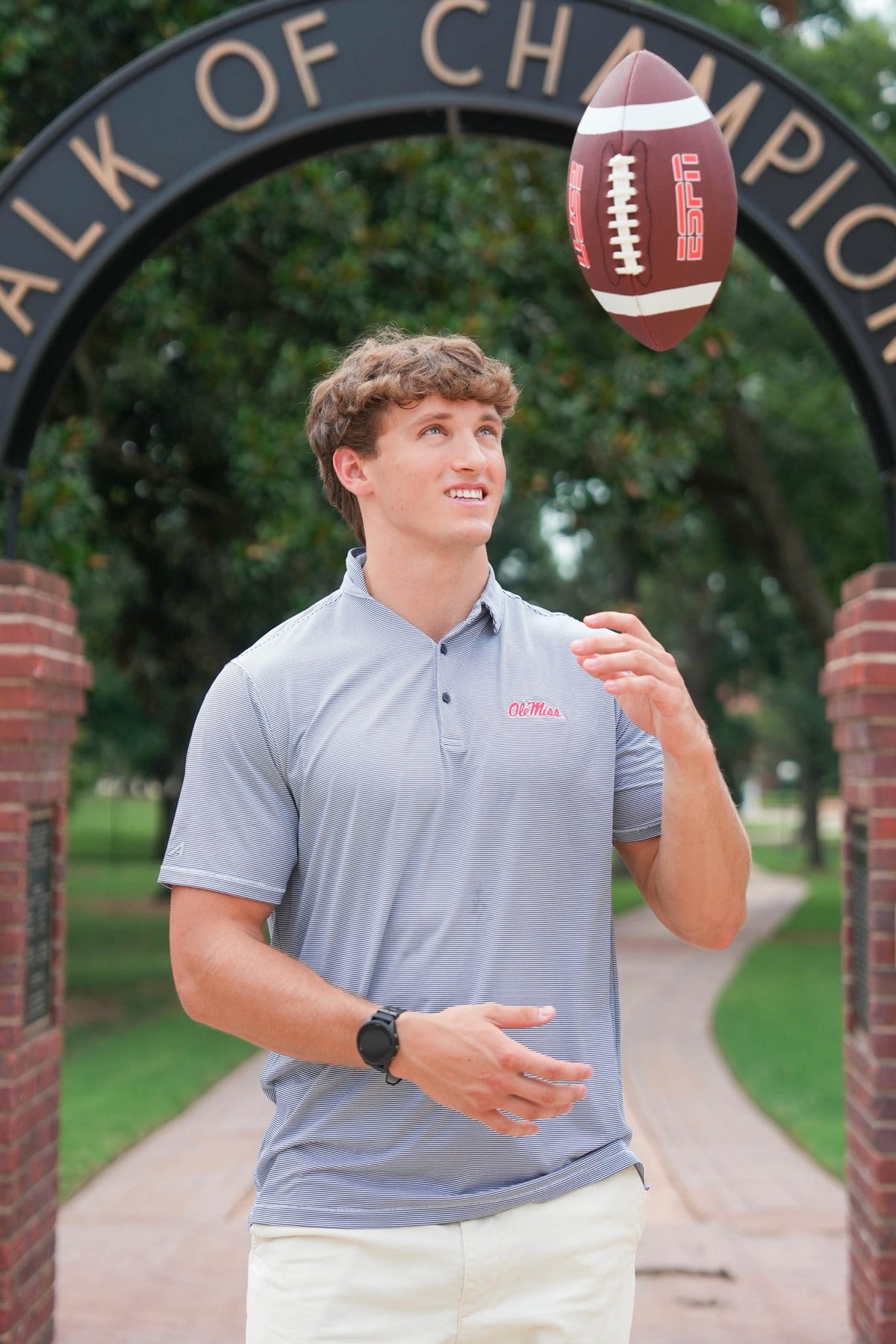 A man wearing a light blue polo shirt and white pants stands under a brick archway with a football floating in the air, looking up and smiling. The background shows a park path and trees.