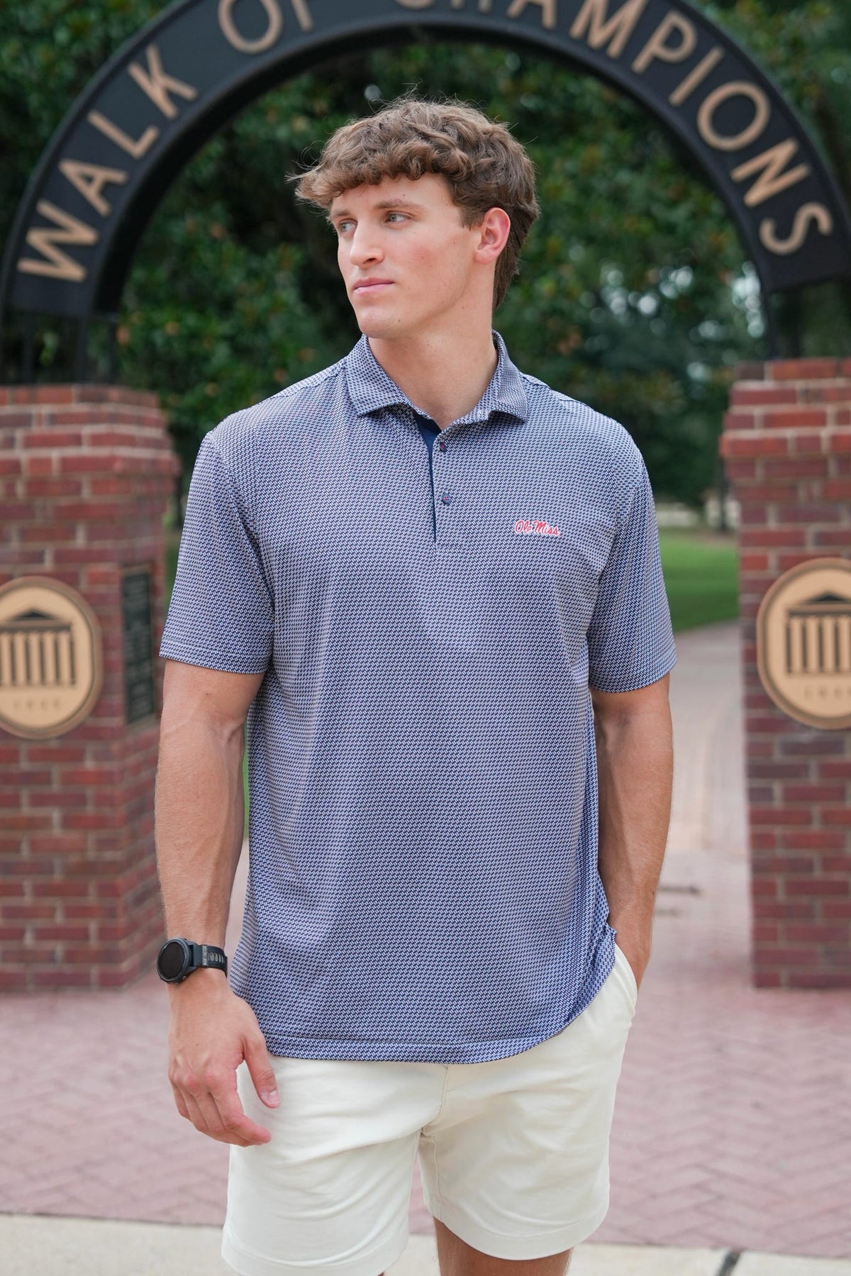 A man wearing a blue and white patterned polo shirt with a small red logo on the chest, paired with white shorts, standing in front of a brick archway with the words 'WALK OF CHAMPIONS'.