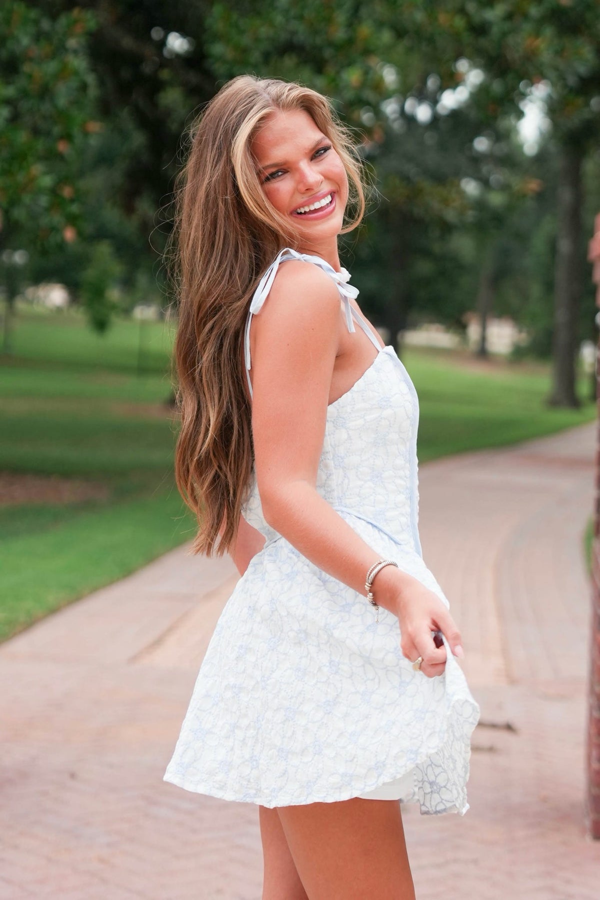 A woman with long brown hair stands on a brick pathway in a park, wearing a white sleeveless mini dress with a subtle floral pattern.