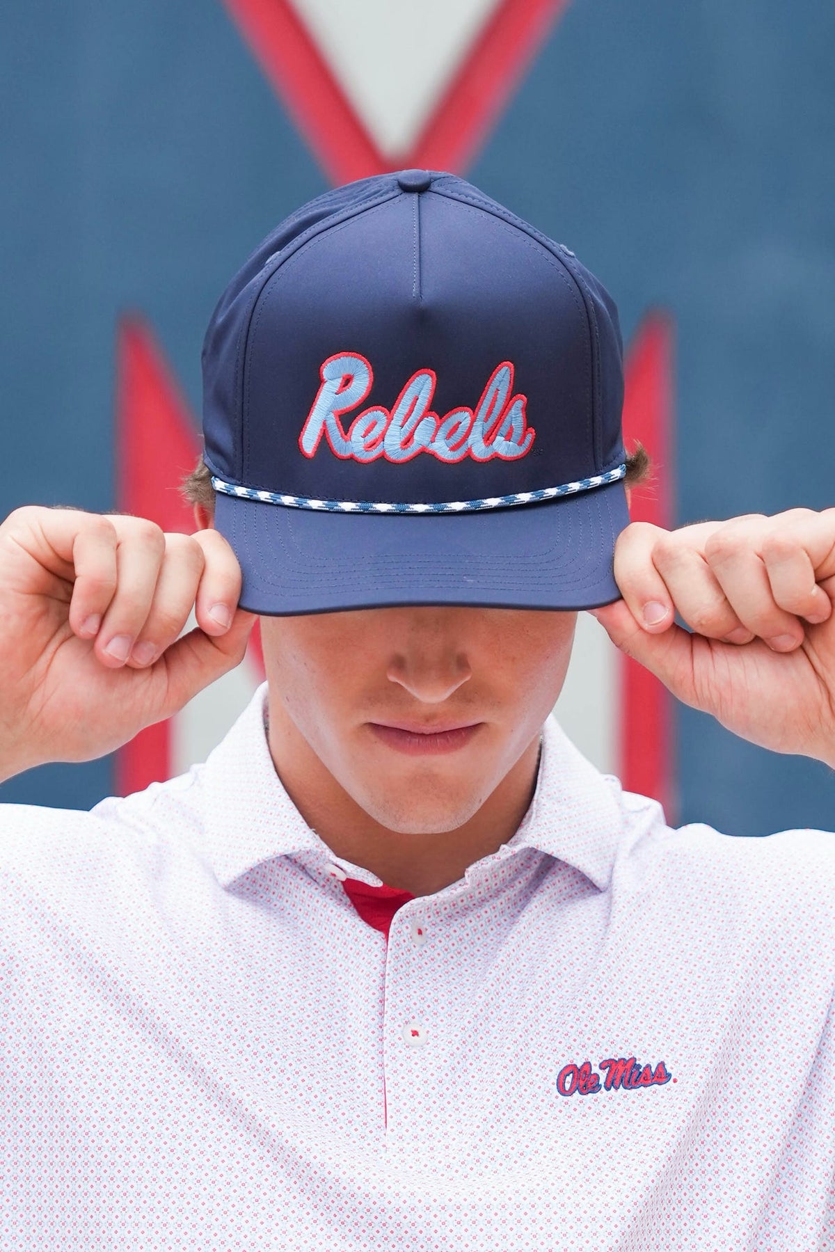 A man wearing a navy blue snapback cap with 'Rebels' embroidered in red and white, holding the brim down over his eyes. He is wearing a white patterned polo shirt with an 'Ole Miss' logo on the chest, set against a blue and red background.