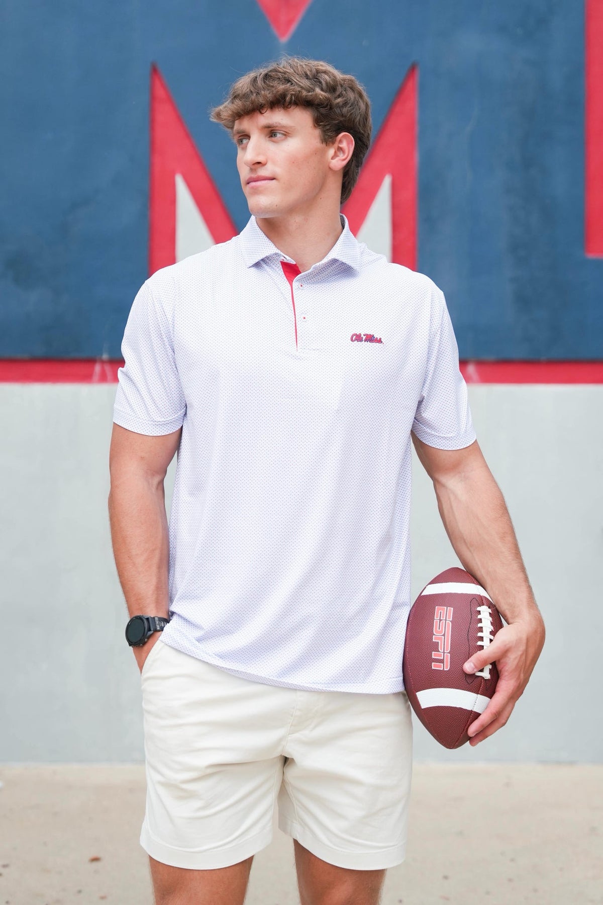 A man wearing a white short-sleeve polo shirt with a red inner collar and a small red logo on the left chest, paired with cream shorts and holding a football, standing in front of a blue and red wall.