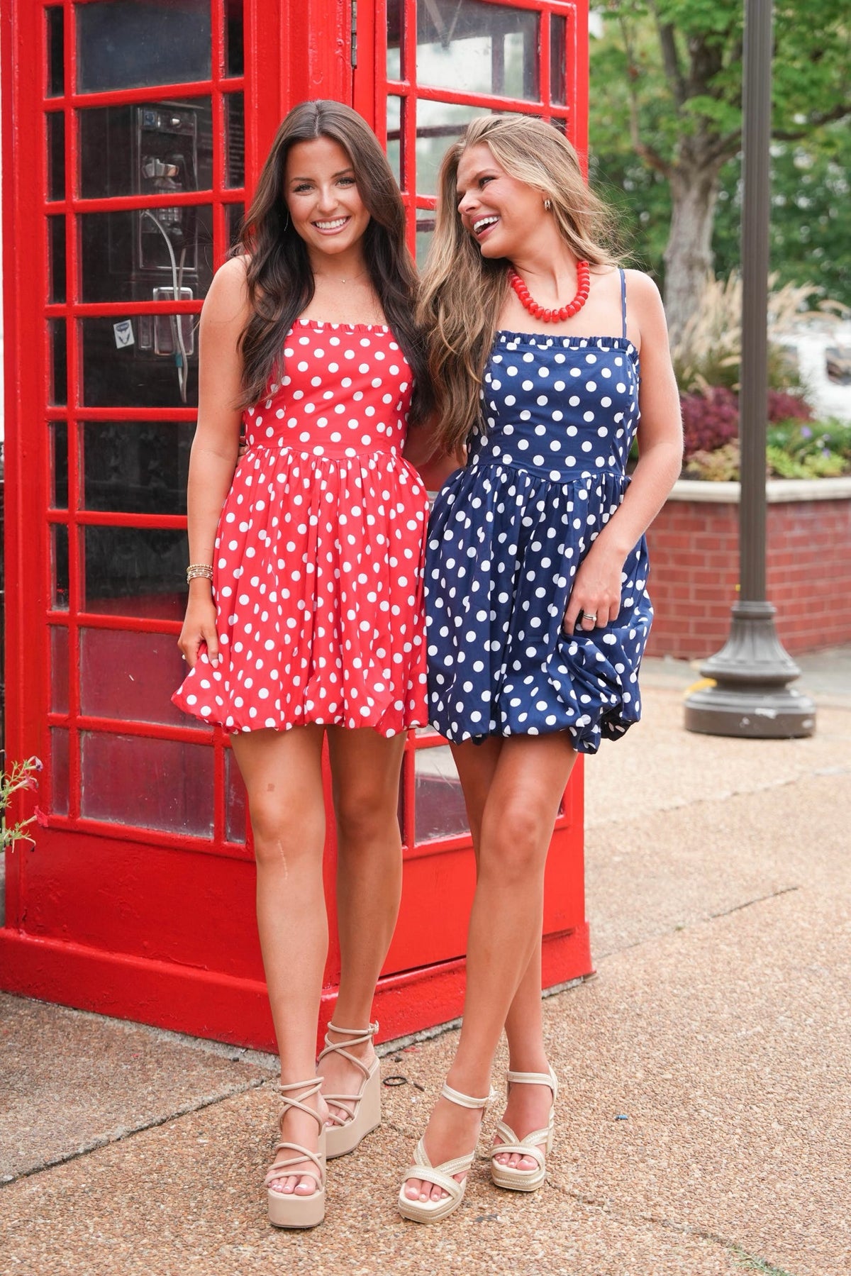 Two women standing outdoors in front of a red telephone booth, wearing matching strapless polka-dot mini dresses. One dress is red with white polka dots, and the other is navy blue with white polka dots.