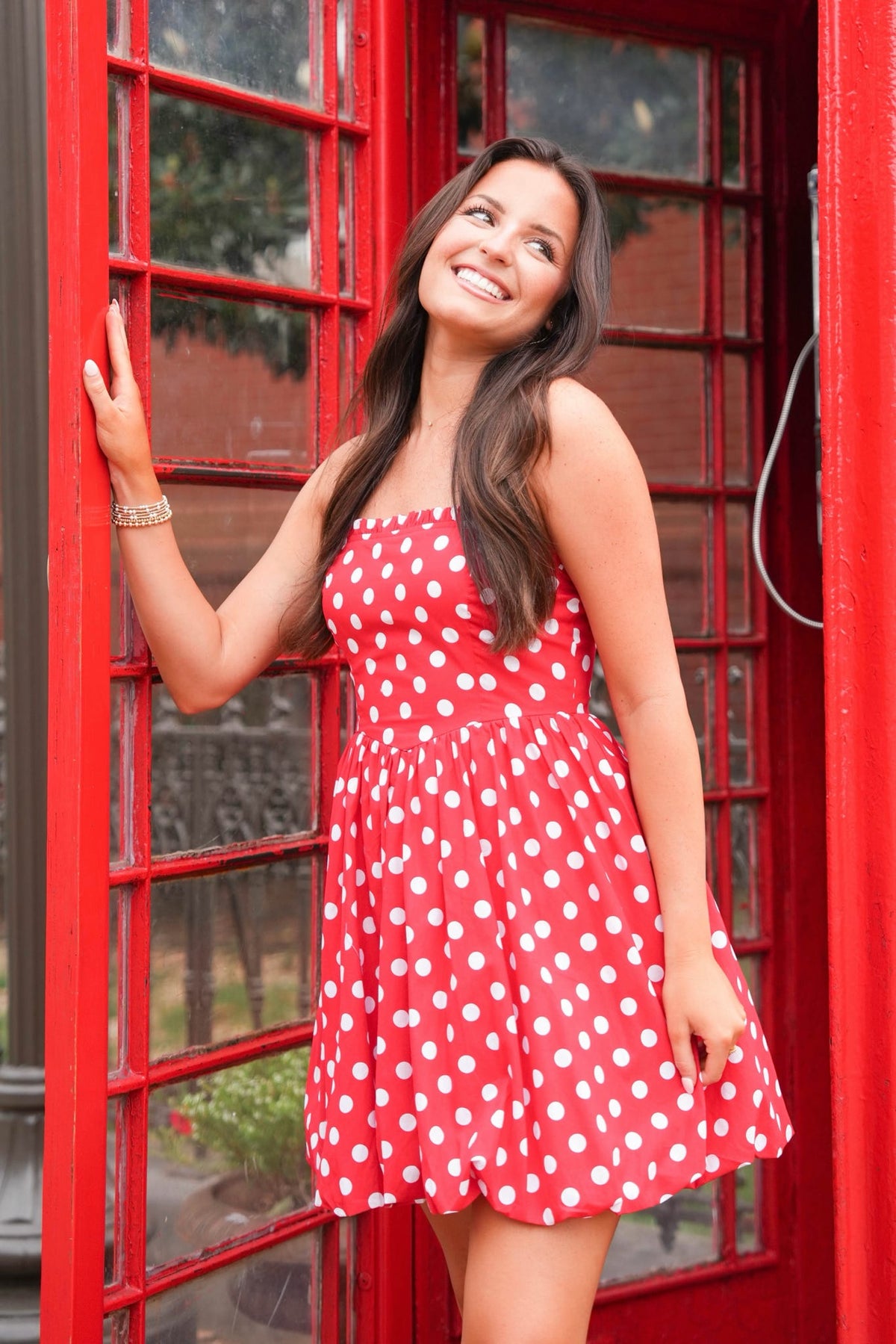A woman wearing a red strapless dress with white polka dots stands smiling next to a classic red telephone booth.