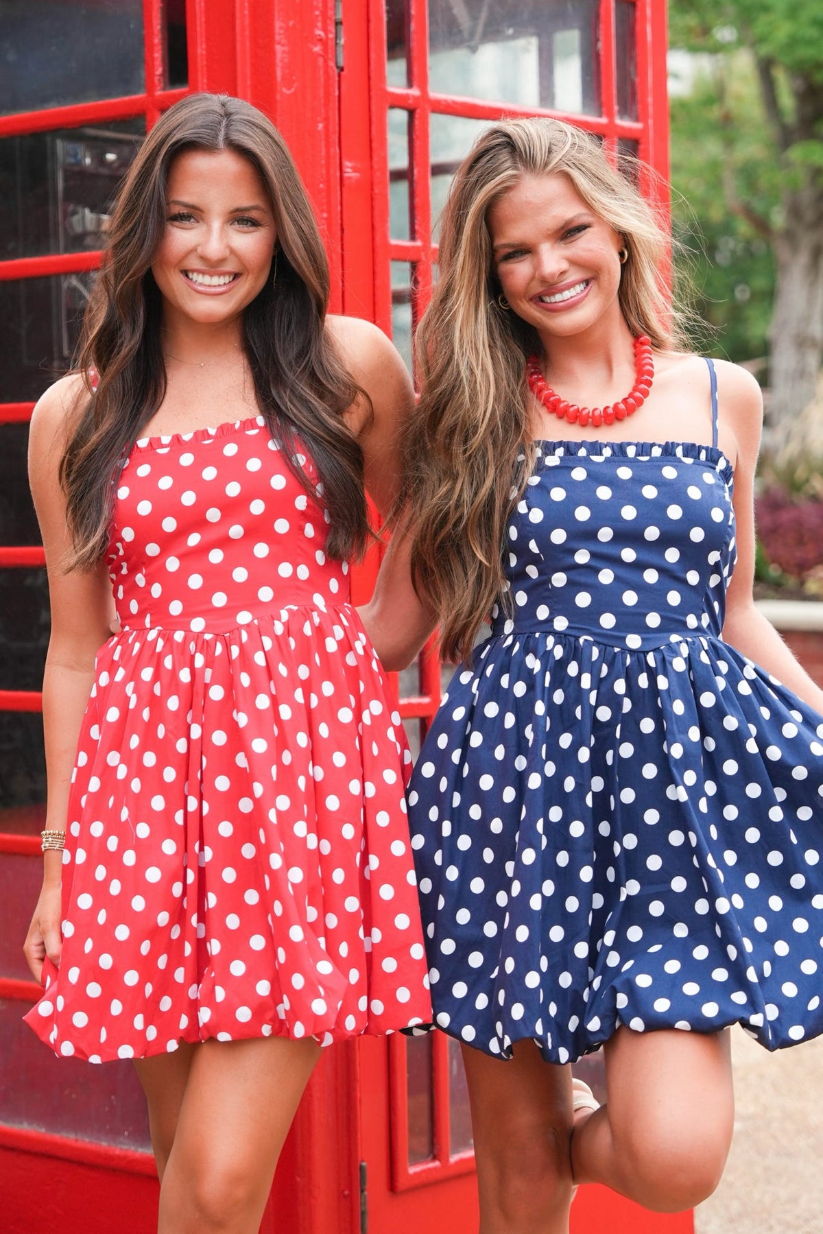 Two women modeling short polka-dot dresses in front of a red telephone booth. One dress is red with white polka dots, and the other is navy blue with white polka dots.
