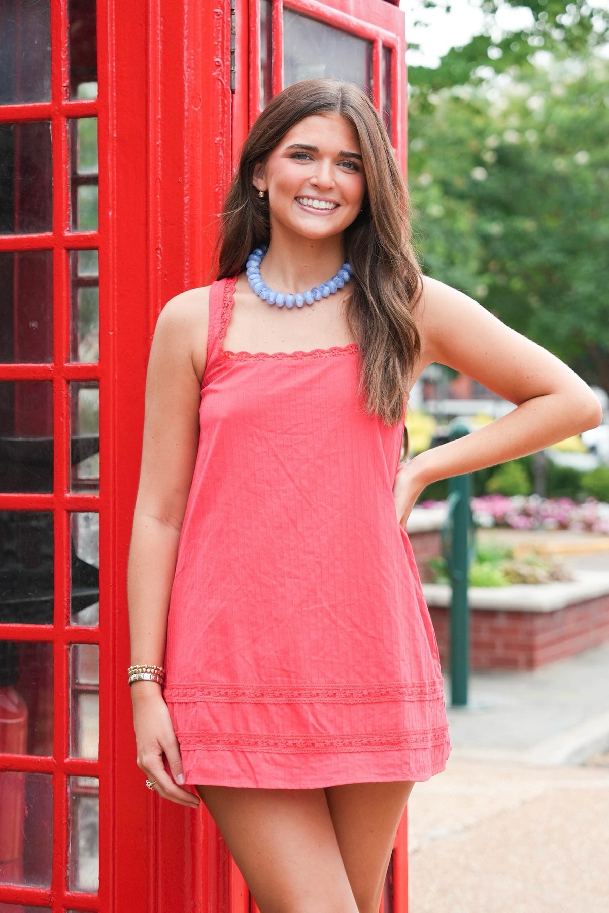 A woman wearing a coral pink sleeveless mini dress with scalloped edges and a lace hem, posing in front of a classic red telephone booth.