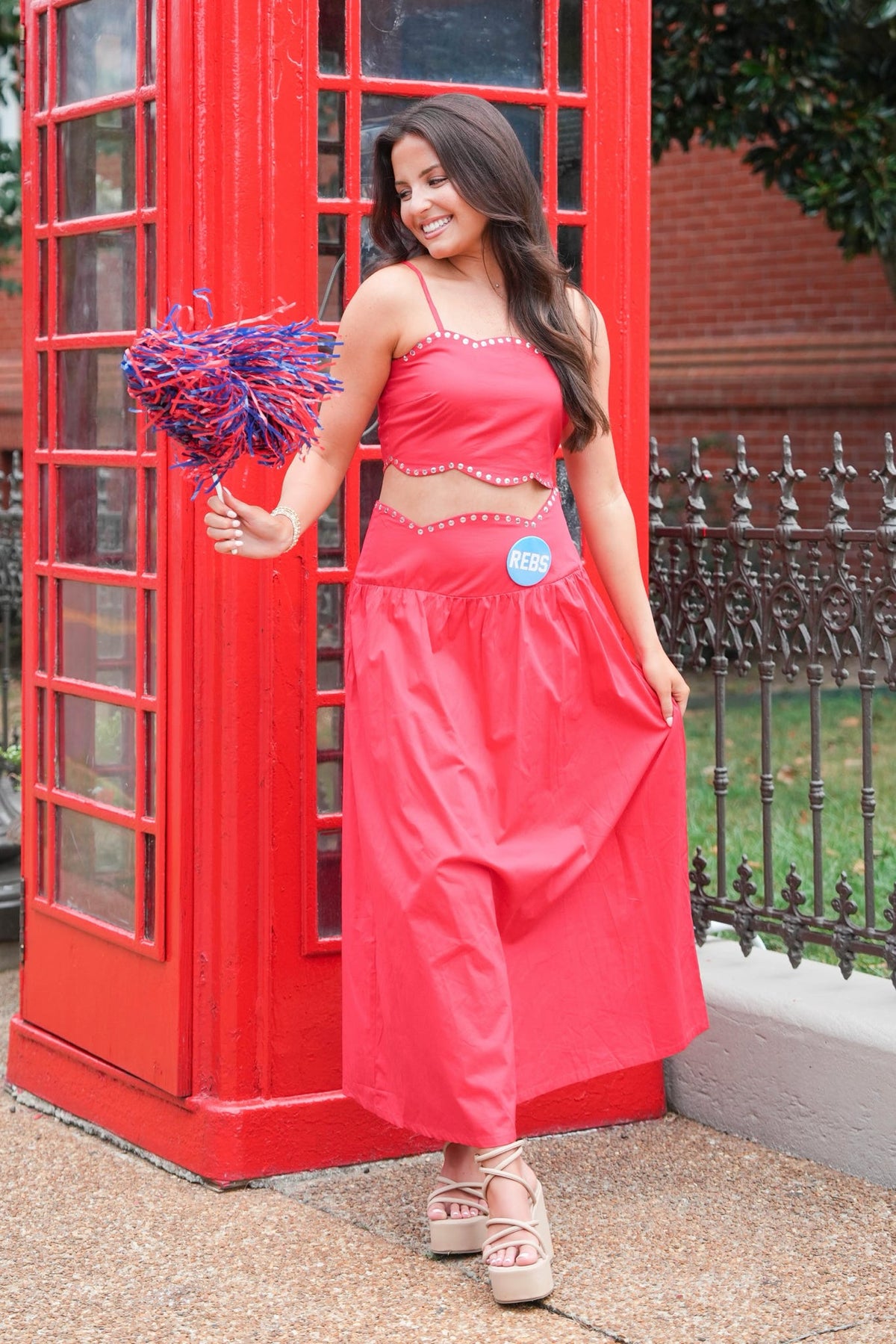 A woman modeling a coral pink two-piece outfit consisting of a halter crop top and matching wide-leg skirt, standing in front of a classic red telephone booth and holding a blue and pink pom-pom.