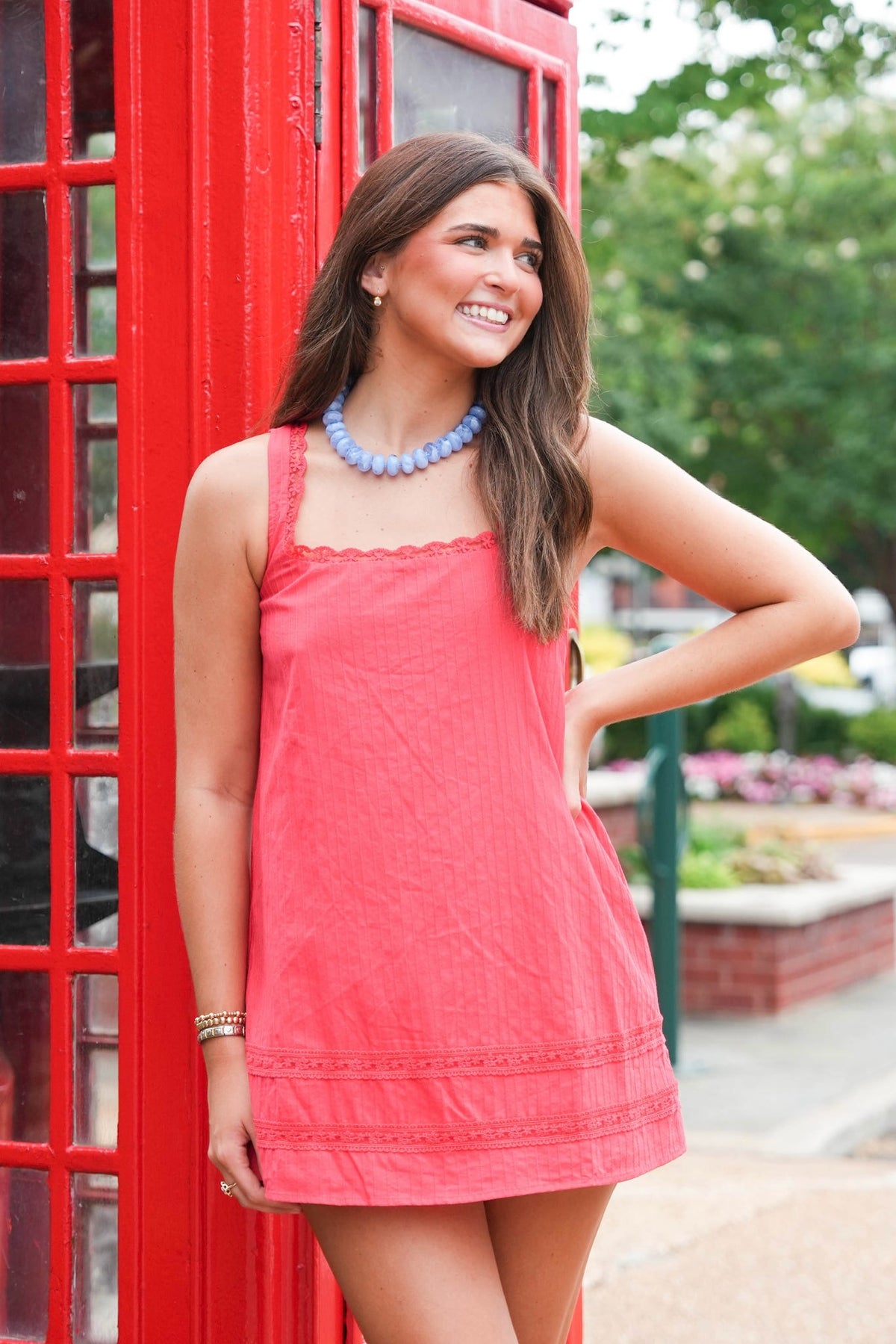 A woman wearing a coral pink sleeveless mini dress with a scalloped neckline and embroidered hem, standing next to a classic red telephone booth.
