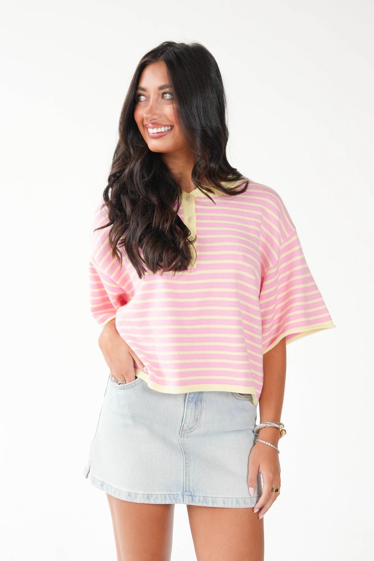 A woman modeling a pink and white striped short-sleeve top with yellow trim, paired with a light blue denim mini skirt, against a plain white background.