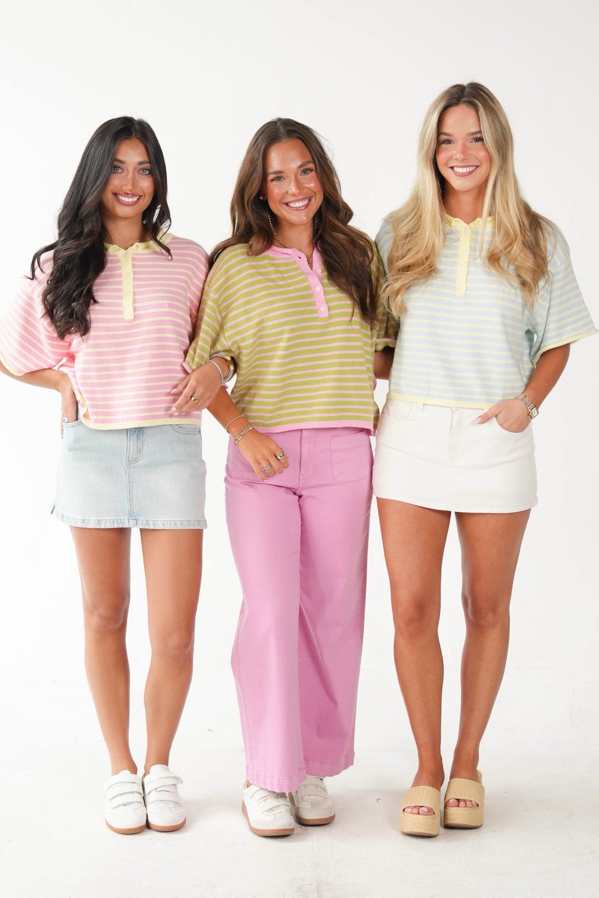 Three women standing side-by-side modeling pastel striped short-sleeve polo shirts. The shirt on the left is pink and white, paired with a denim mini skirt and white sneakers.
