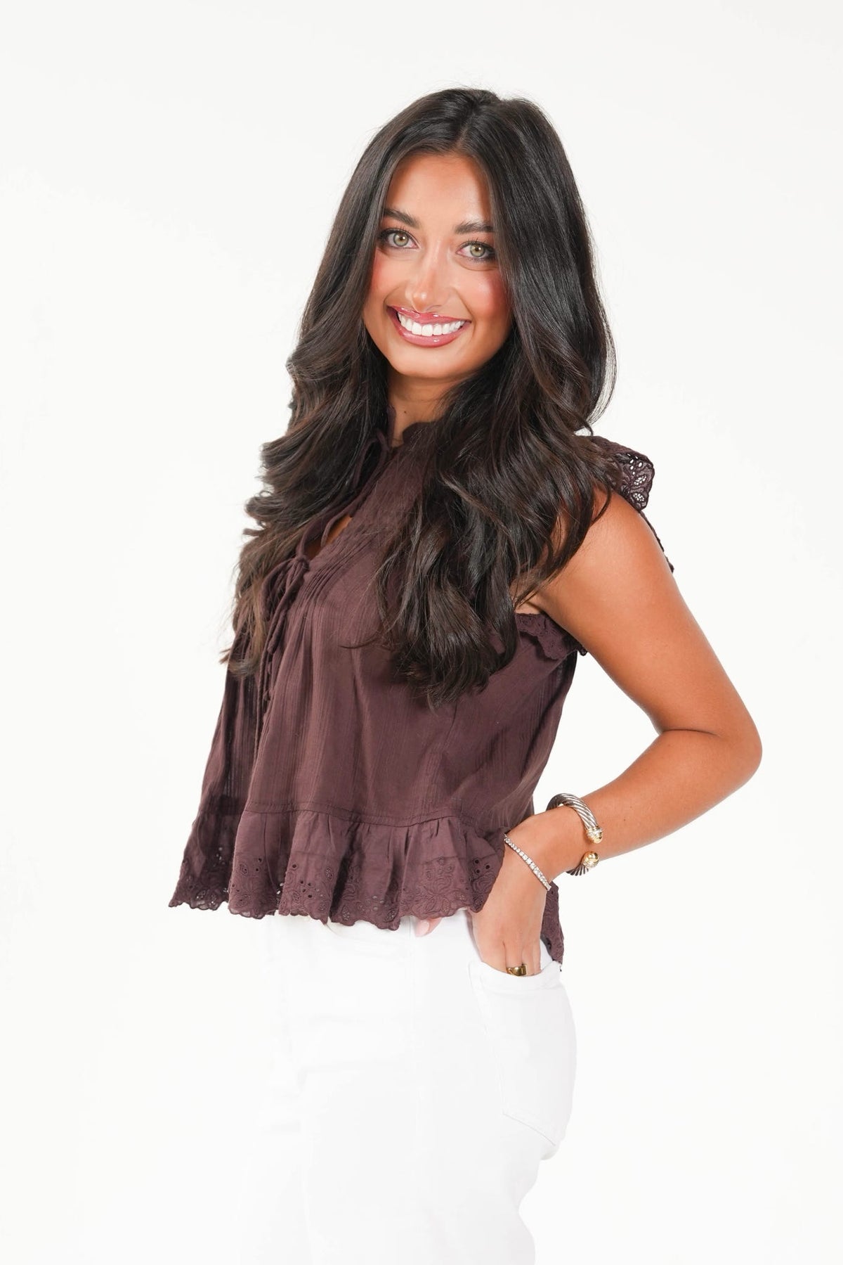 A smiling woman with long dark hair wearing a sleeveless, ruffled dark brown top and white pants, standing against a plain white background.
