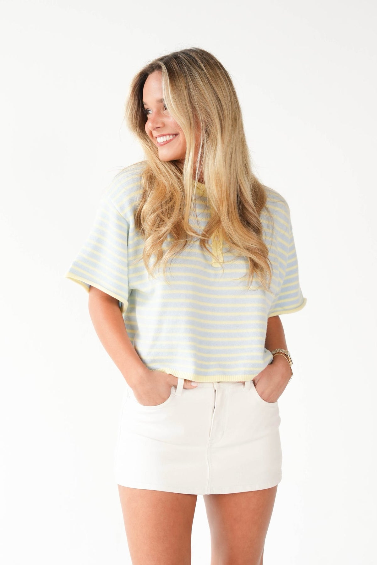 A woman with long blonde hair is shown from the waist up, wearing a light blue and yellow striped short-sleeve t-shirt and white shorts against a plain white background.